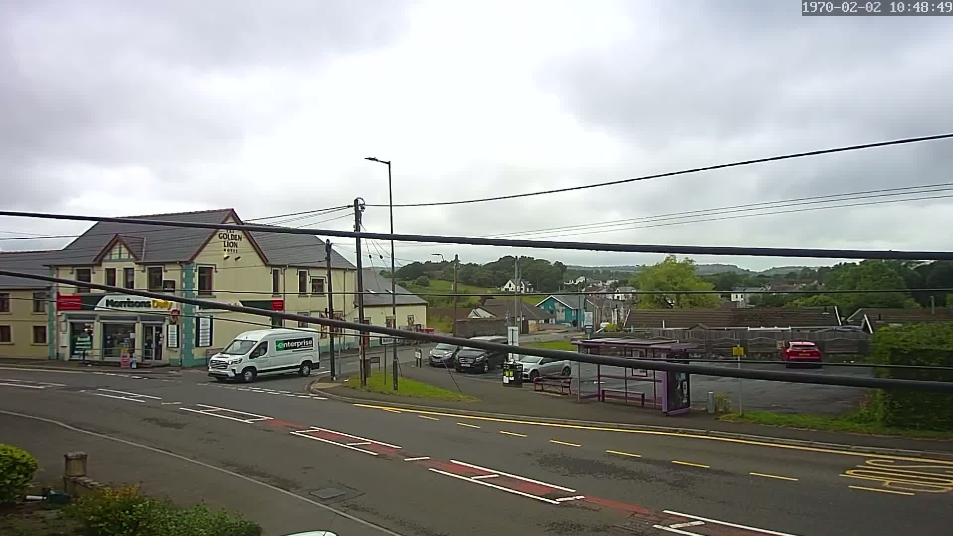 A street scene on an overcast day shows a hotel, a small shop, several cars parked, a bus shelter, and a van driving away from the camera.