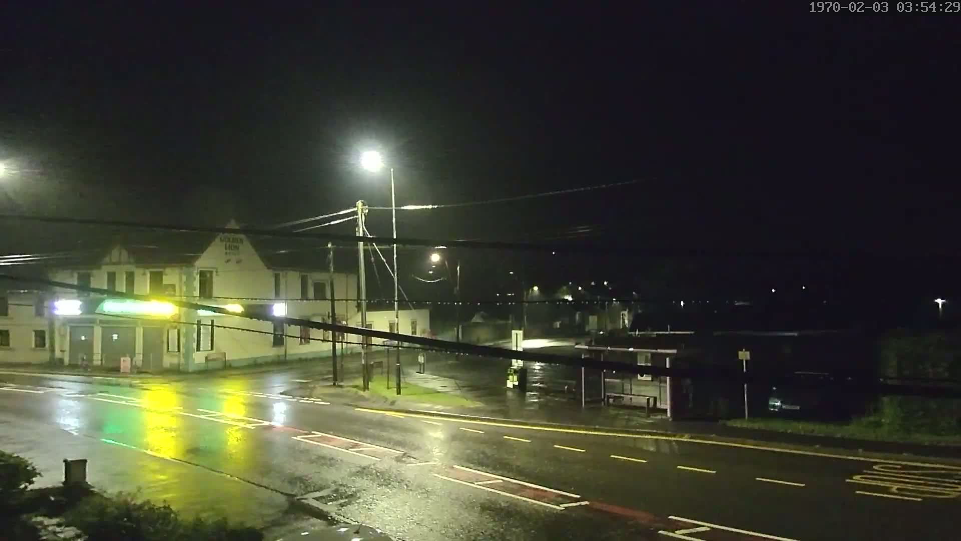 A wet, dark street at night shows a building with lights, a bus stop, and streetlights illuminating the rain-slicked road.