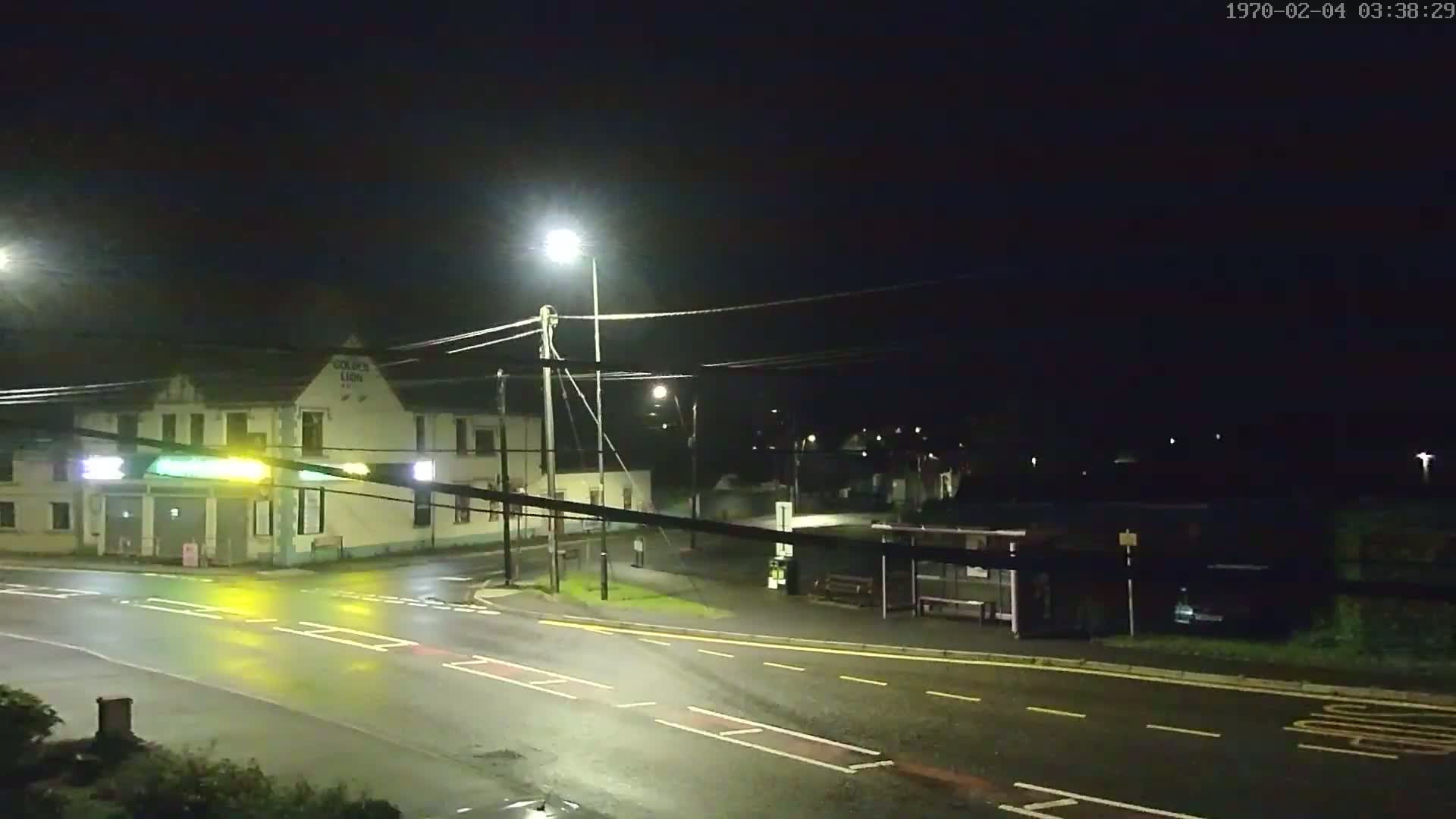 A nighttime street scene shows a wet road intersecting with another road near a building with streetlights illuminating the area.