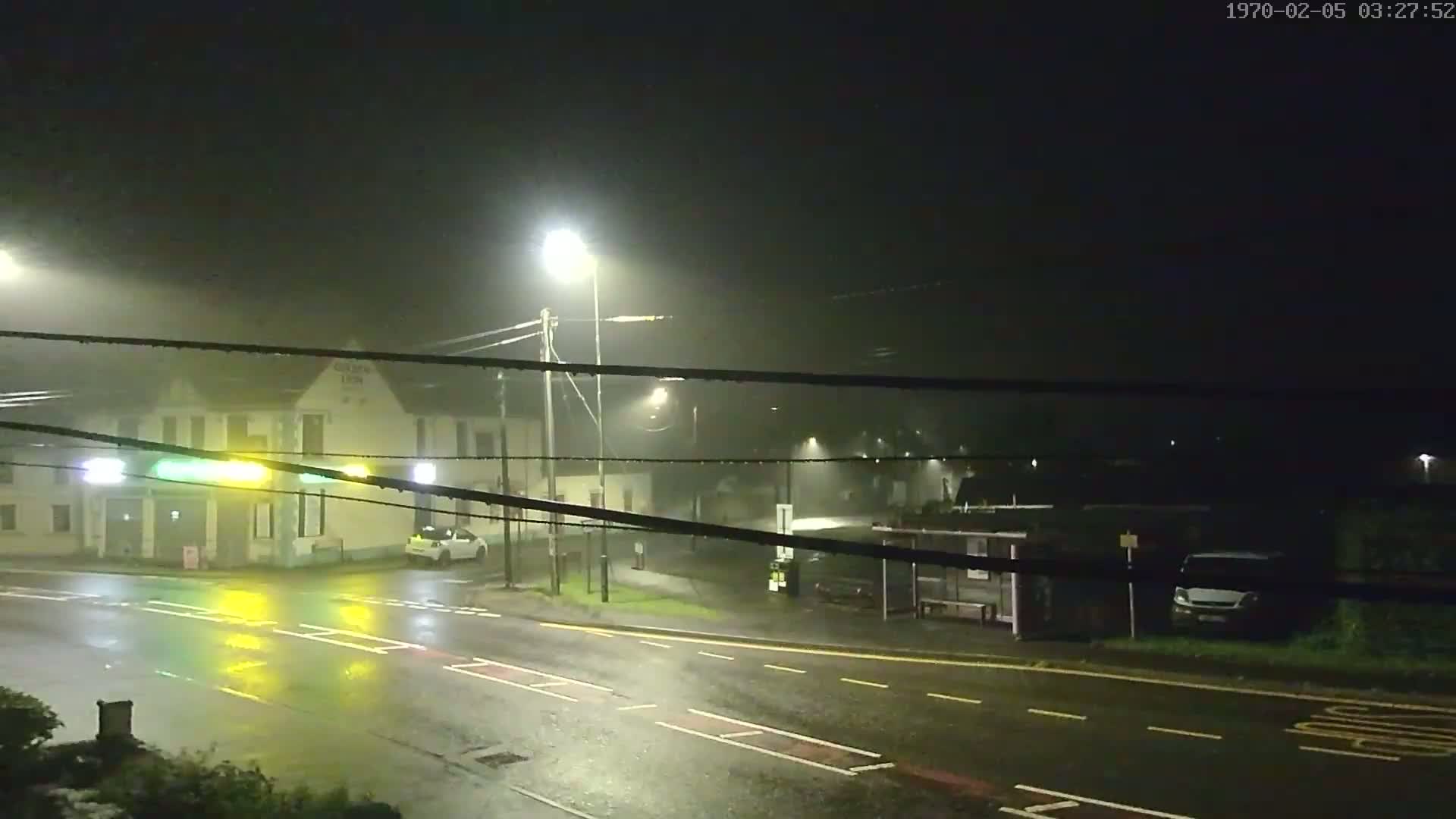 A rain-slicked street at night, illuminated by streetlights, shows a building, a bus stop, and several vehicles under foggy conditions.
