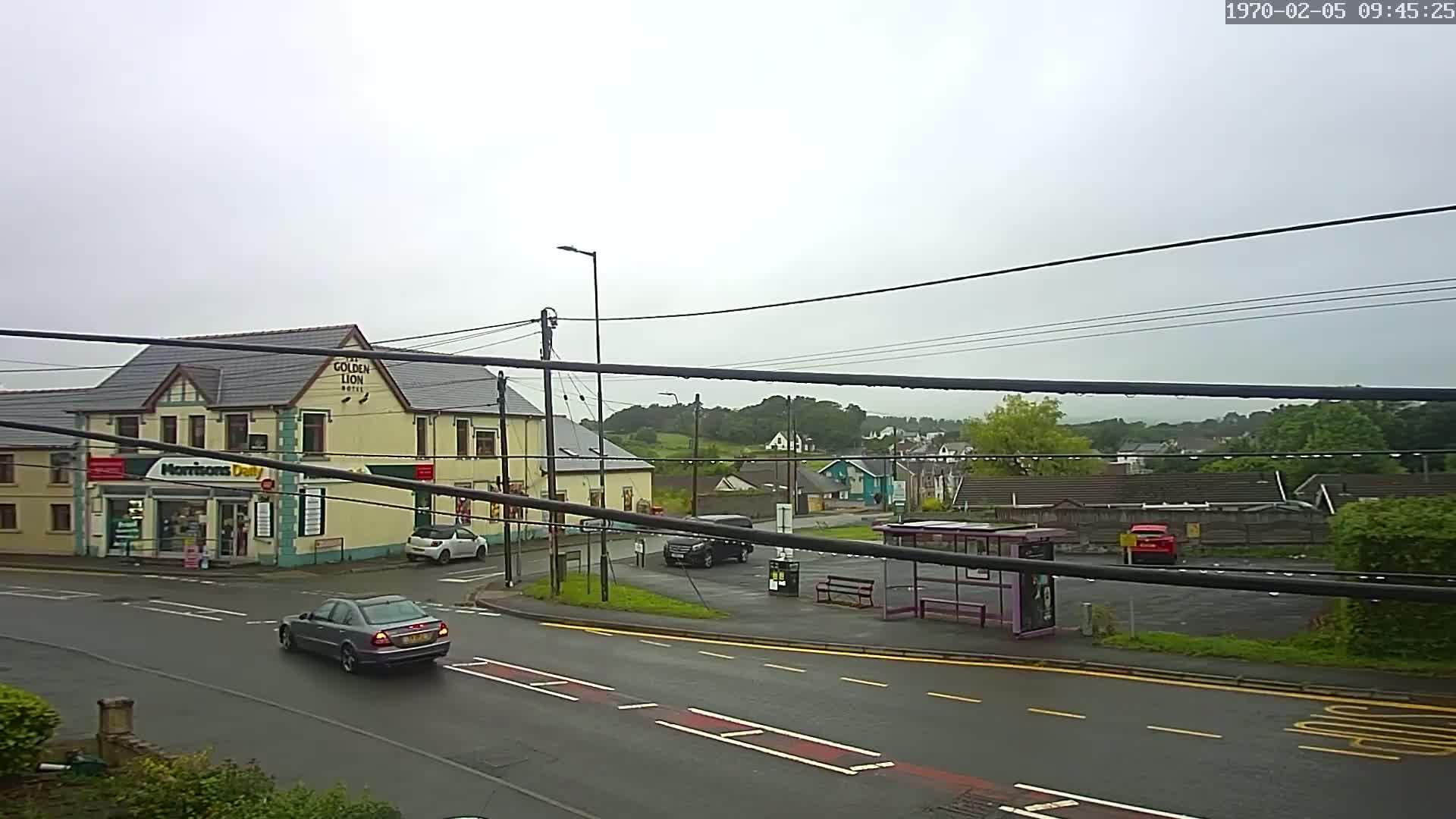 A wet, overcast day reveals a street scene with cars, a bus stop, a small shop, and a hotel, all situated near a small, hilly area.
