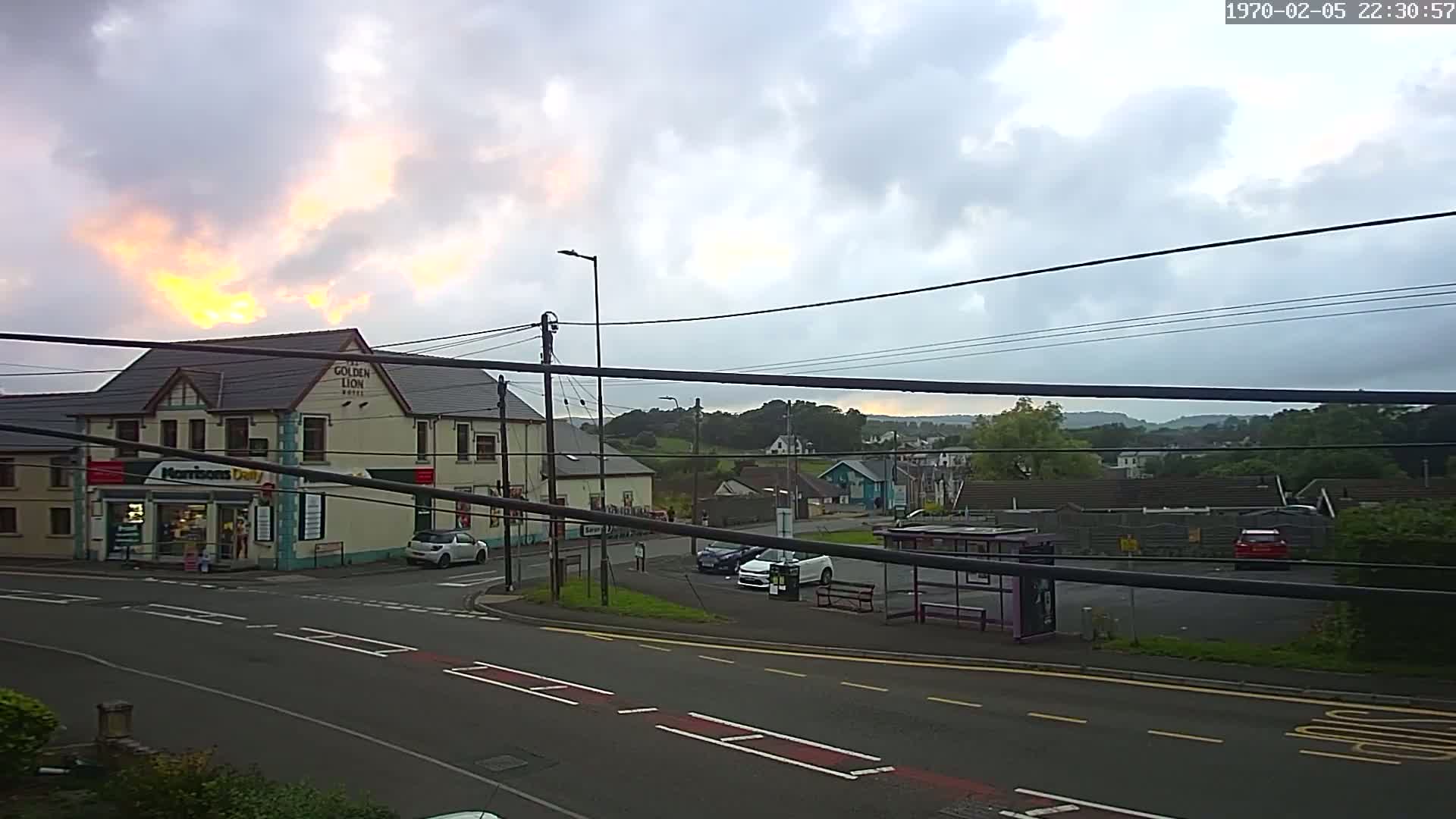 A street scene under a cloudy sky with a setting sun, showing buildings, cars, and a bus stop.