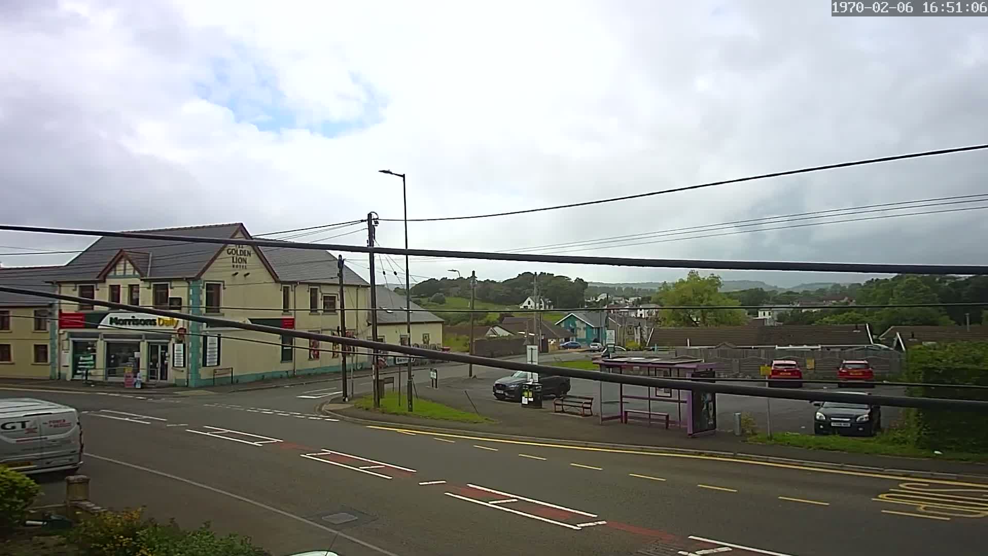 A street scene on an overcast day shows a crossroads with a bus stop, a small shop, a hotel, and several parked cars.