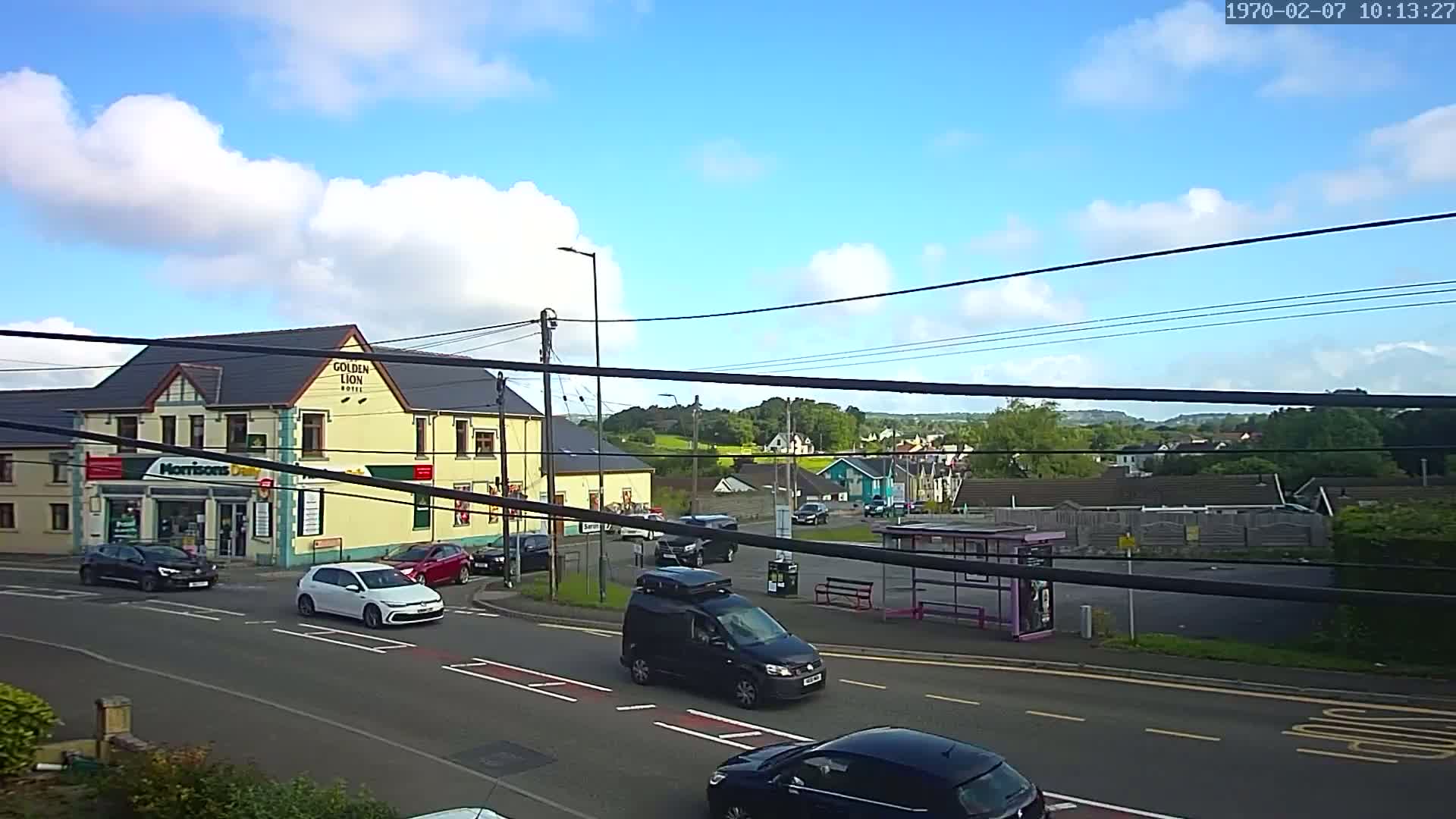 A partly sunny day reveals a street scene with cars at an intersection near a yellow hotel and a small shop, with a bus stop and residential buildings visible in the background.