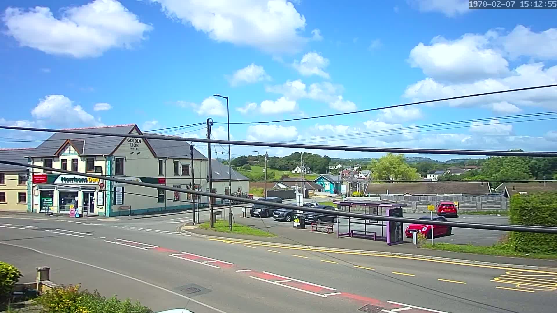 A partly sunny day reveals a street intersection with a pub, a small shop, a bus stop, and several parked cars, all situated in front of a hilly landscape.