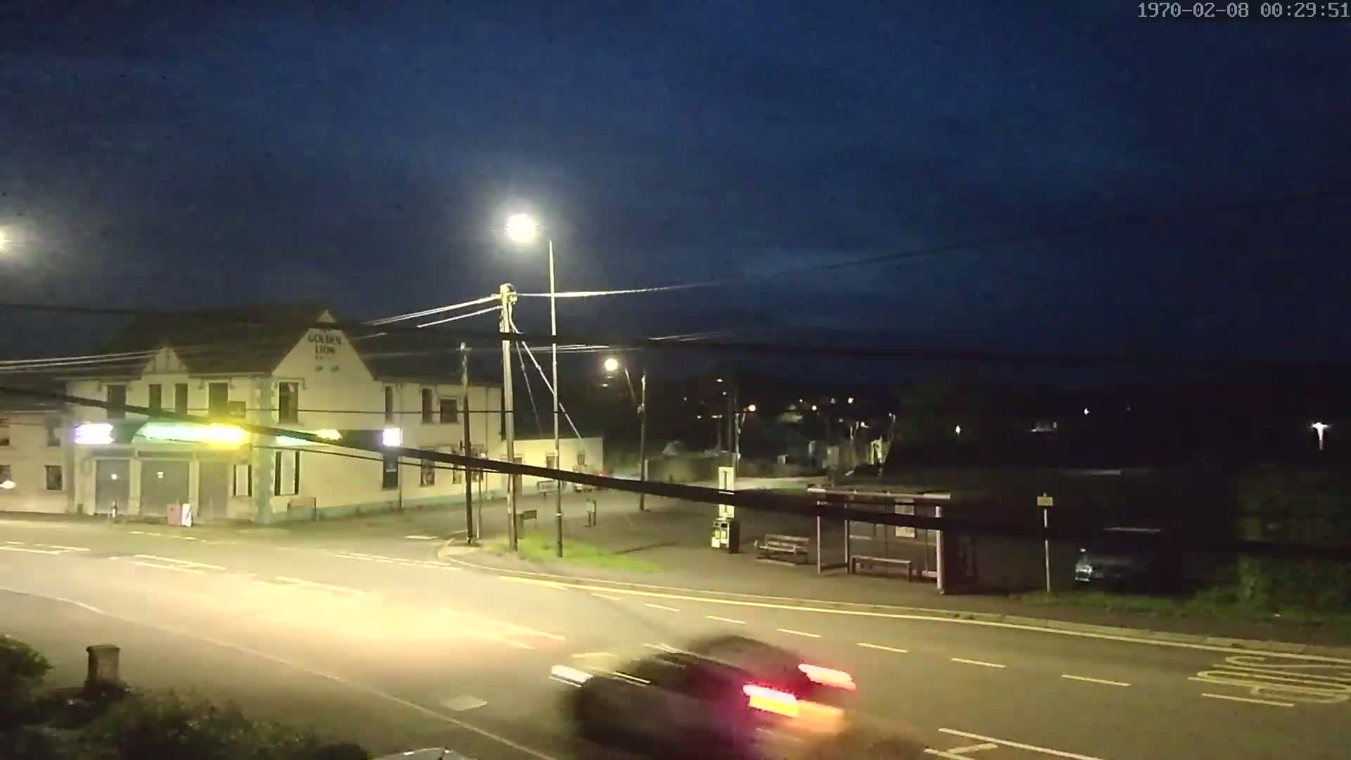 A nighttime street scene shows a car passing a building with lights on, a bus shelter, and streetlights illuminating a mostly empty intersection under a dark sky.