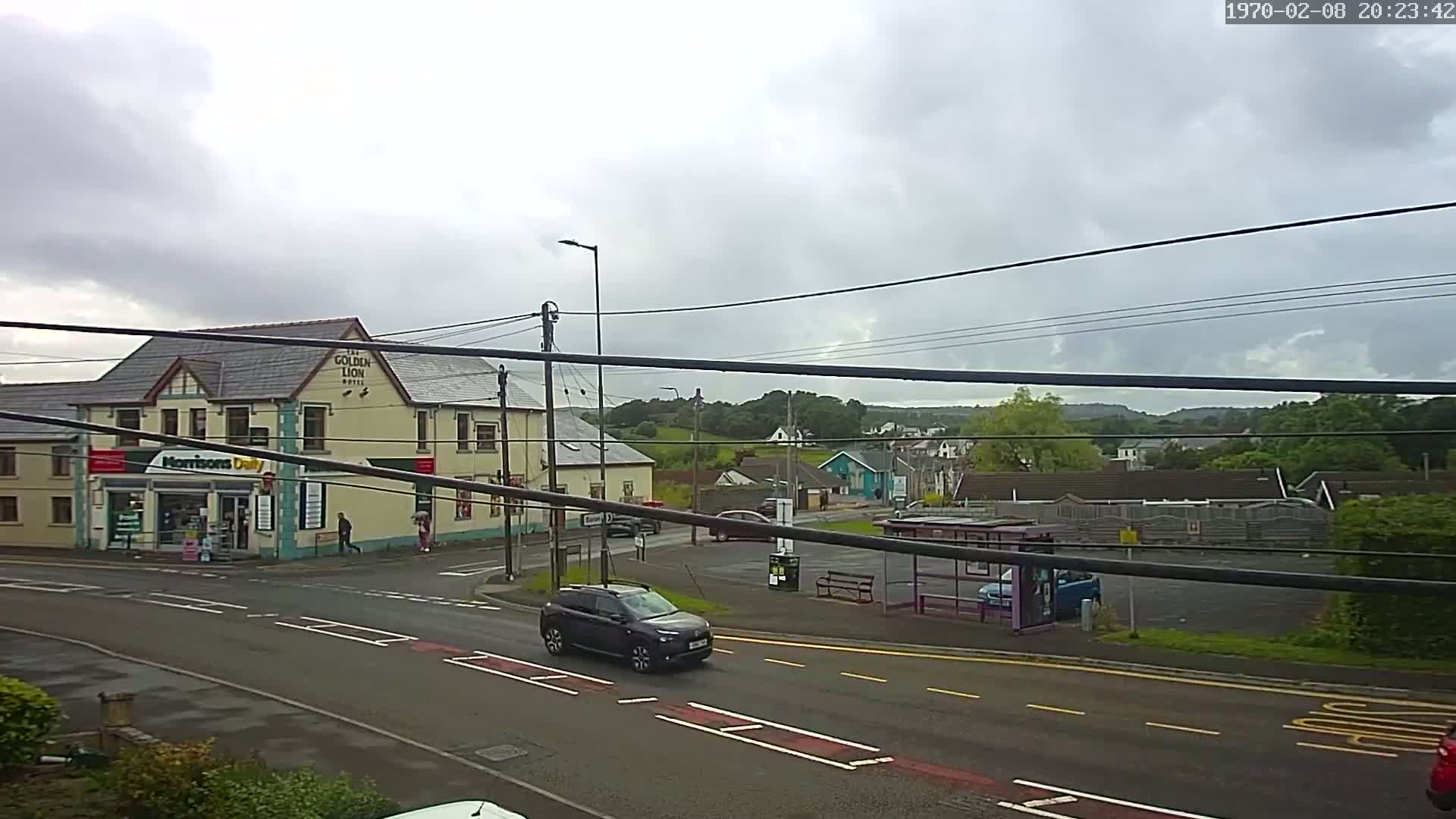A dark-colored SUV drives past a bus stop and a corner store next to a hotel under an overcast sky.