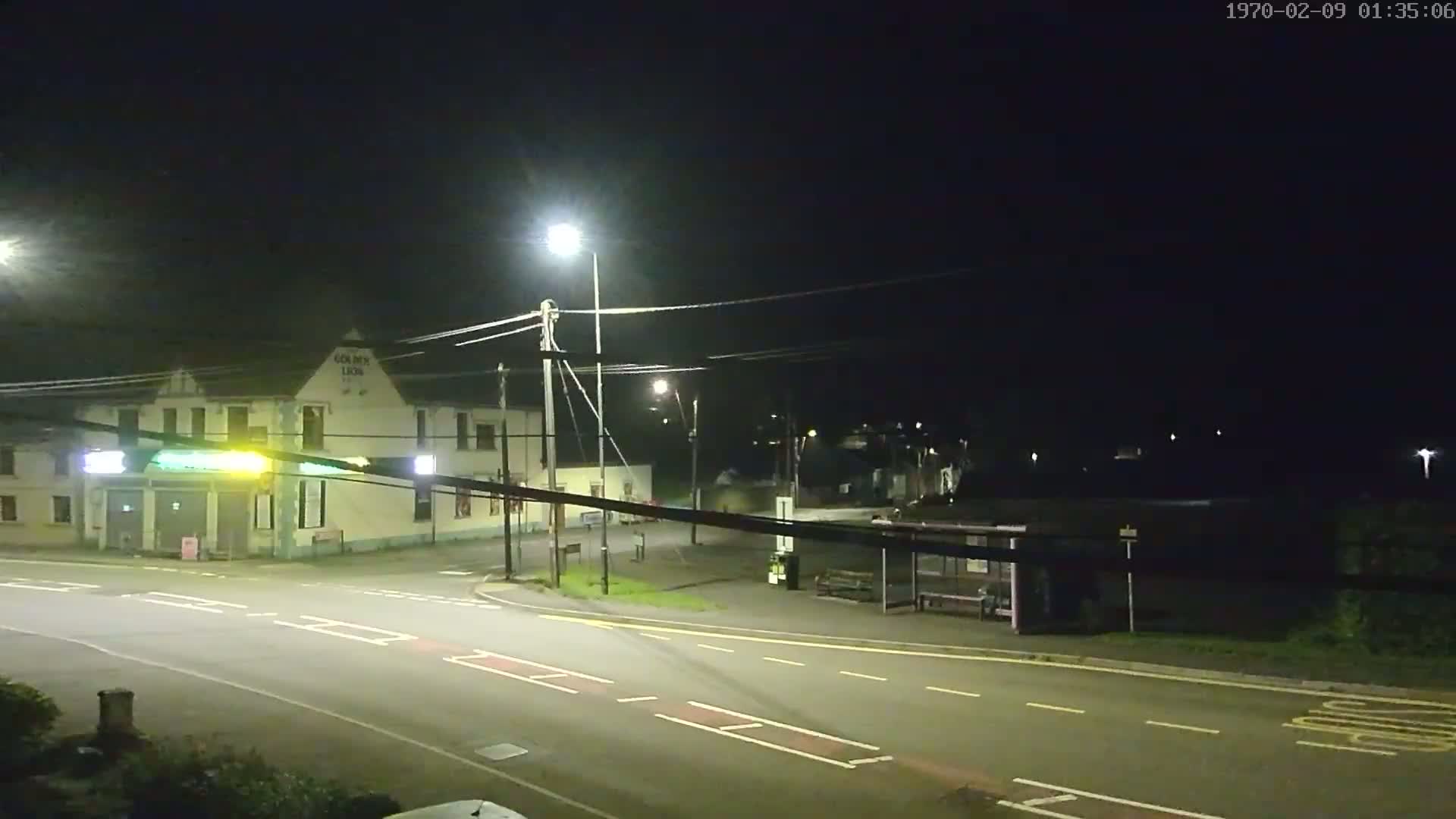 A nighttime street scene shows a building illuminated by streetlights, a bus stop, and a roadway intersection under a clear, dark sky.