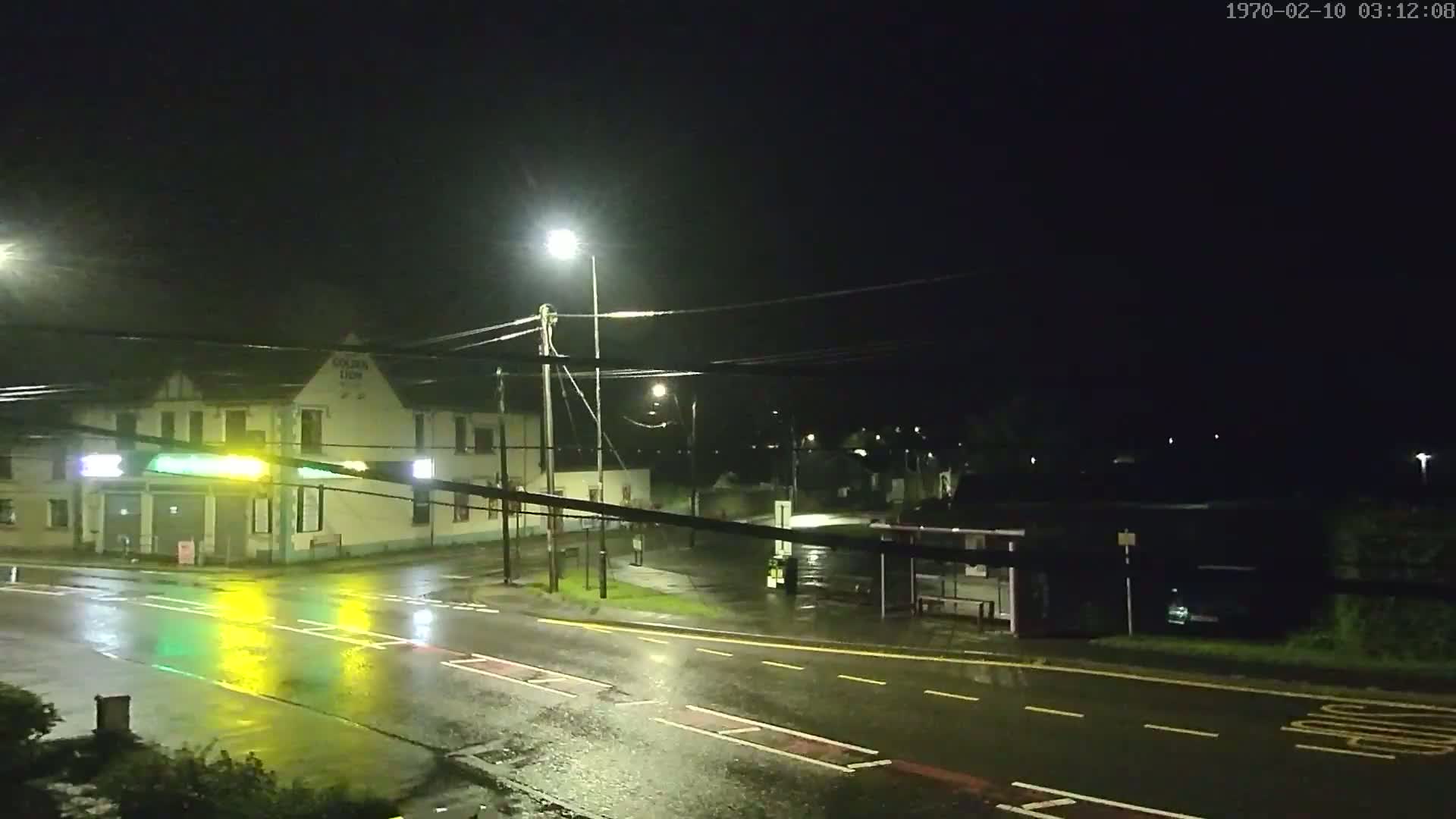 A rain-slicked street intersection at night, illuminated by streetlights, shows a building and a bus stop.