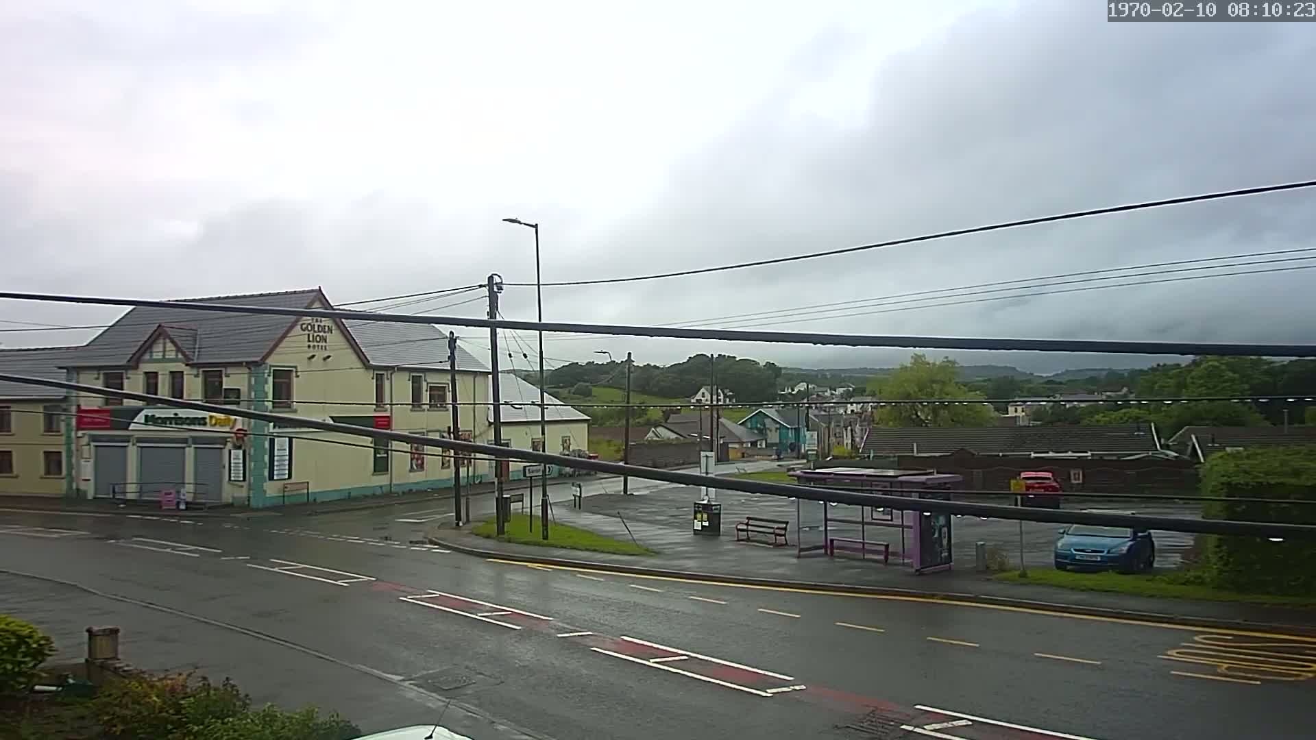 A wet, overcast day reveals a street scene with a light-yellow building, a bus stop, and several cars parked nearby, all situated against a backdrop of green hills.