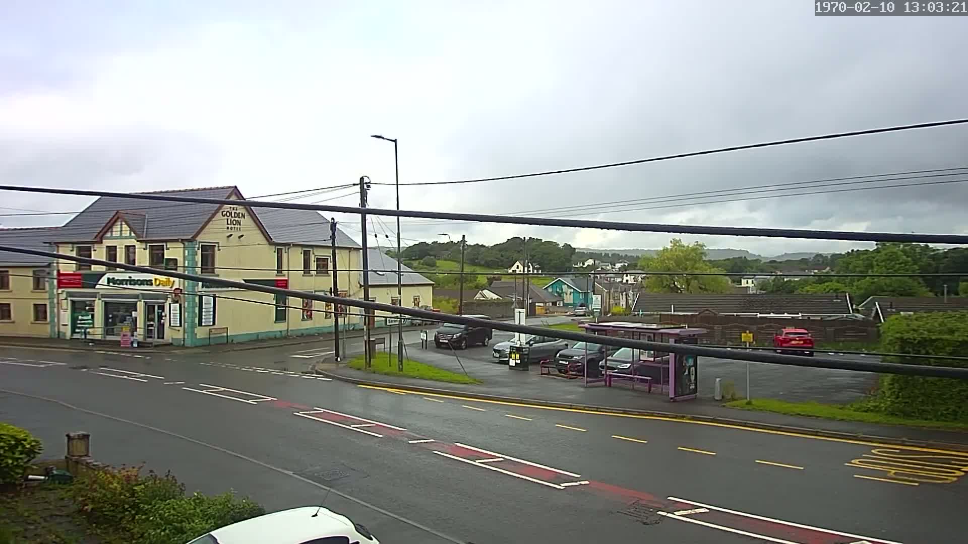 A wet, overcast day reveals a street scene with a yellow building, a small convenience store, parked cars, a bus stop, and a glimpse of a hilly, tree-lined landscape in the distance.