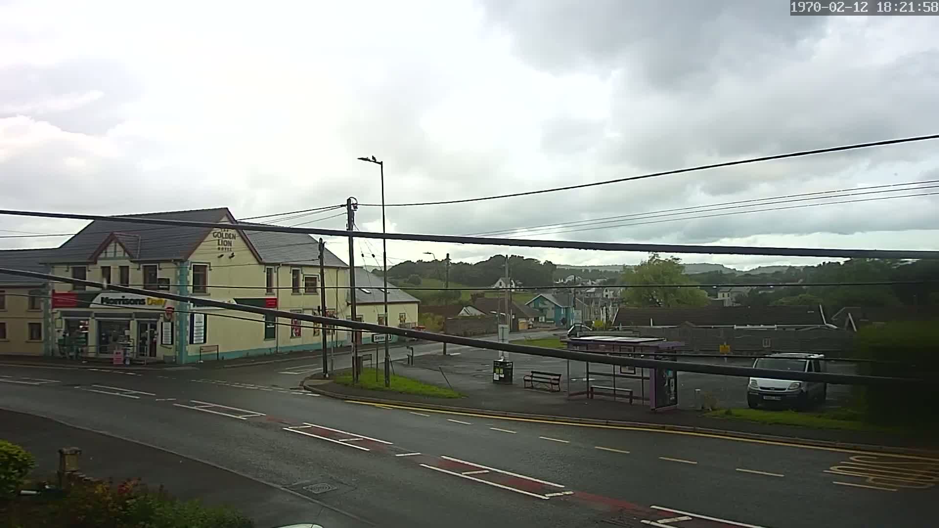 A wet road leads to a corner where a pub and a small shop are located, with a bus shelter and a van parked nearby under a cloudy sky.