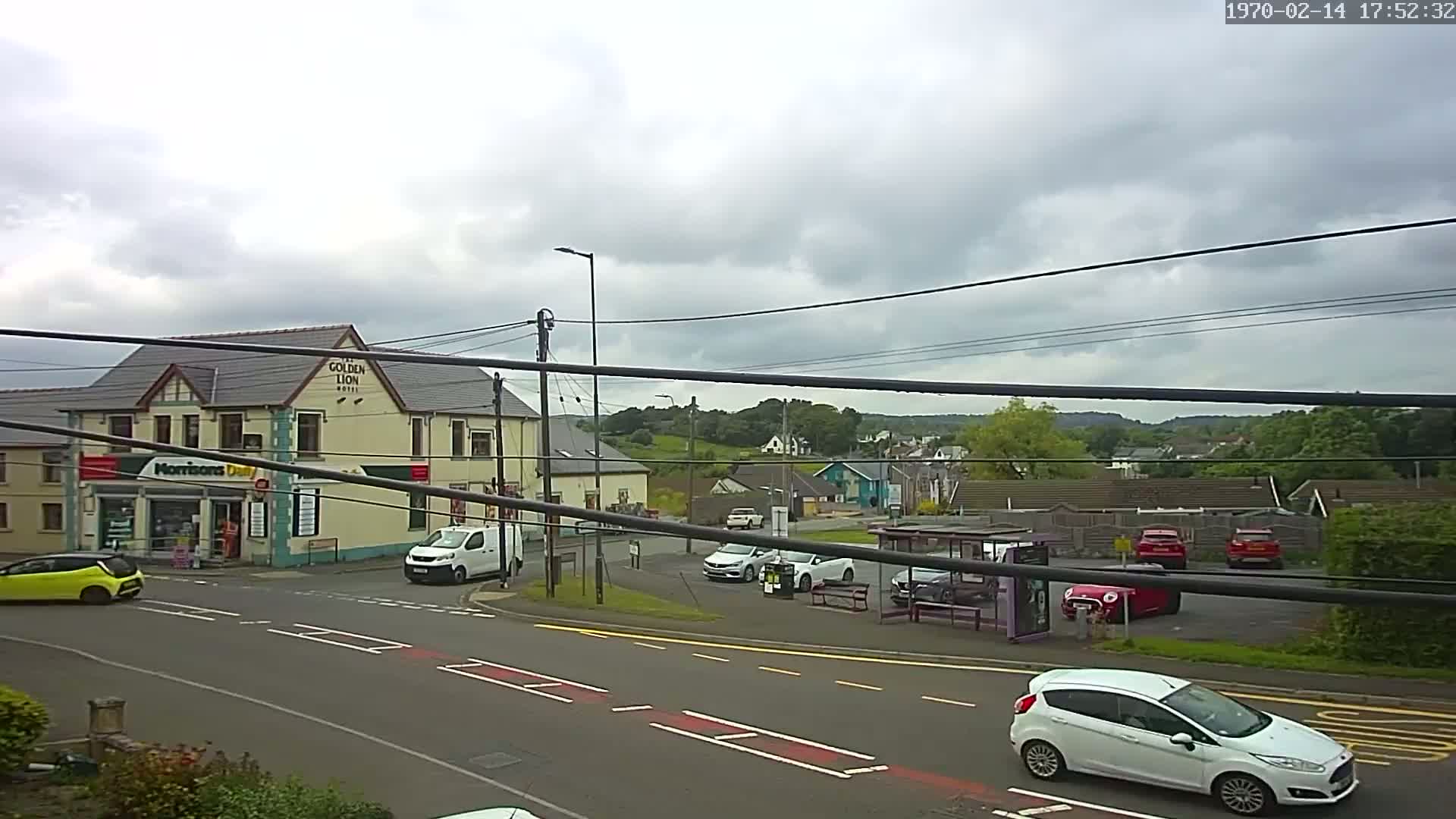 A cloudy day reveals a street scene with a pub, a small shop, several cars, a bus stop, and residential buildings in the background.