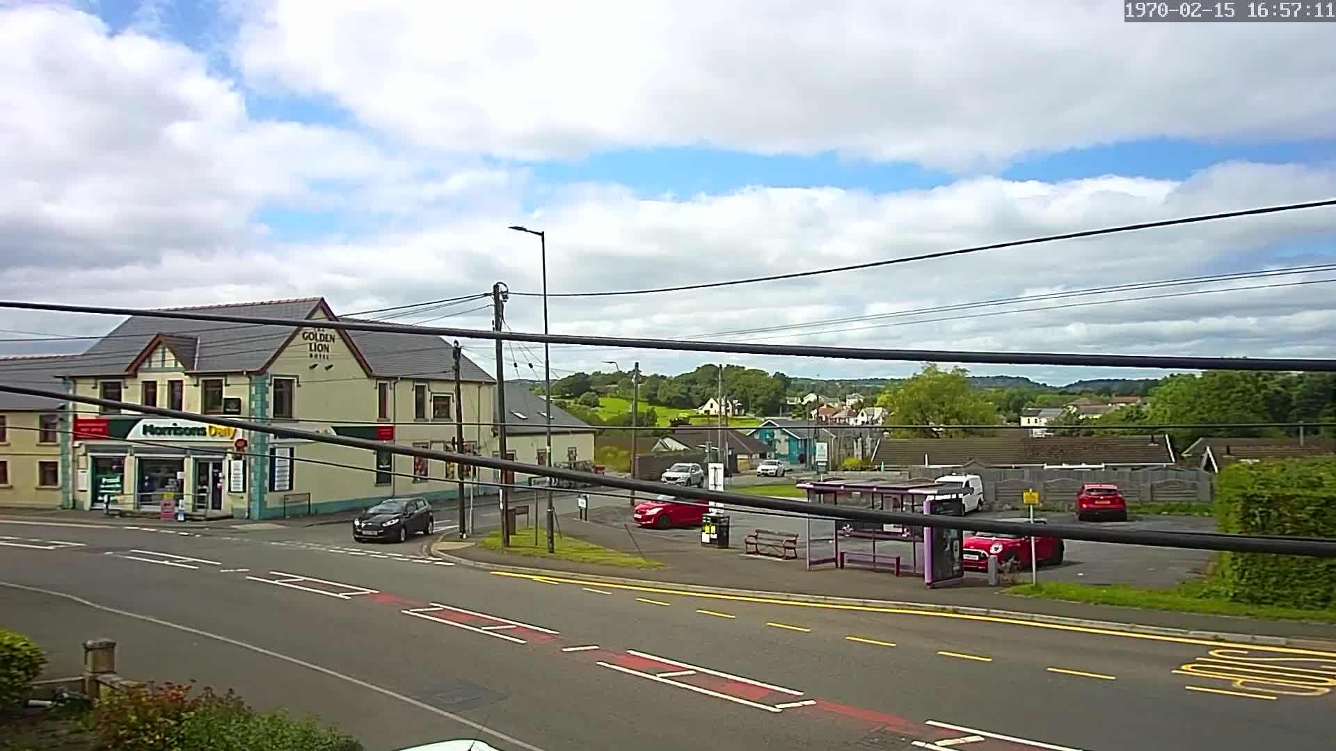 A partly cloudy day reveals a street scene with a pub, a small shop, several cars, and a bus stop, all situated near a residential area with green hills in the background.