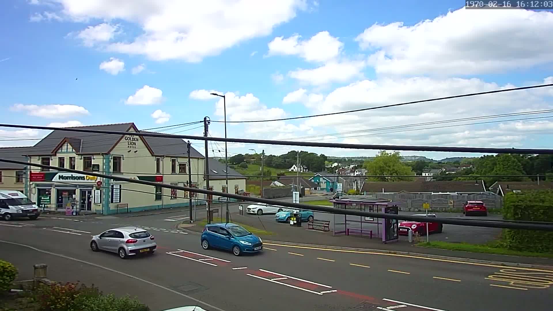 A partly sunny day shows a street scene with several cars, a bus stop, a small market, and a hotel, all situated near a hillside in the background.