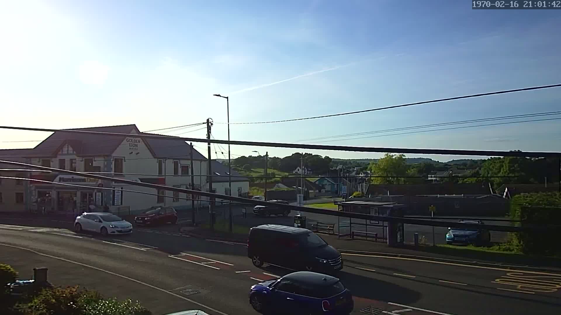 A sunny day reveals a street scene with cars parked and driving past a hotel, other buildings, and greenery in the background.