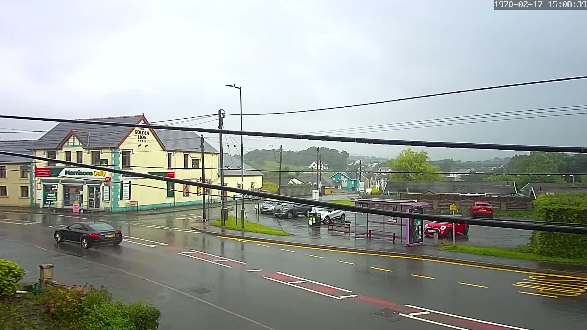 A wet road scene shows a yellow building with a shop and a pub, several cars parked in a lot near a bus stop, and other buildings and greenery visible in the distance under an overcast sky.