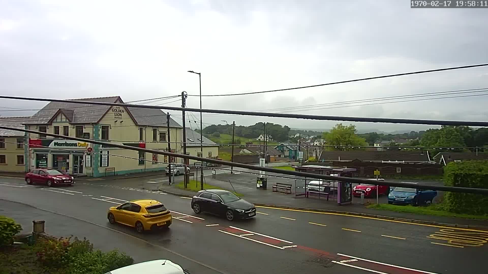 A rain-slicked street scene shows several cars at a crossroads near a pub and convenience store, with a bus stop and residential buildings in the background under a cloudy sky.