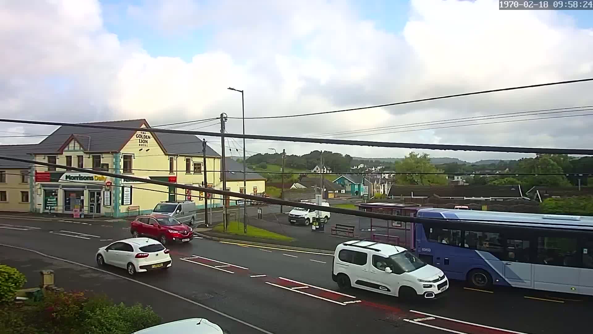Several cars and a bus are driving on a wet road in front of a two-story building and a bus stop, under a cloudy sky.