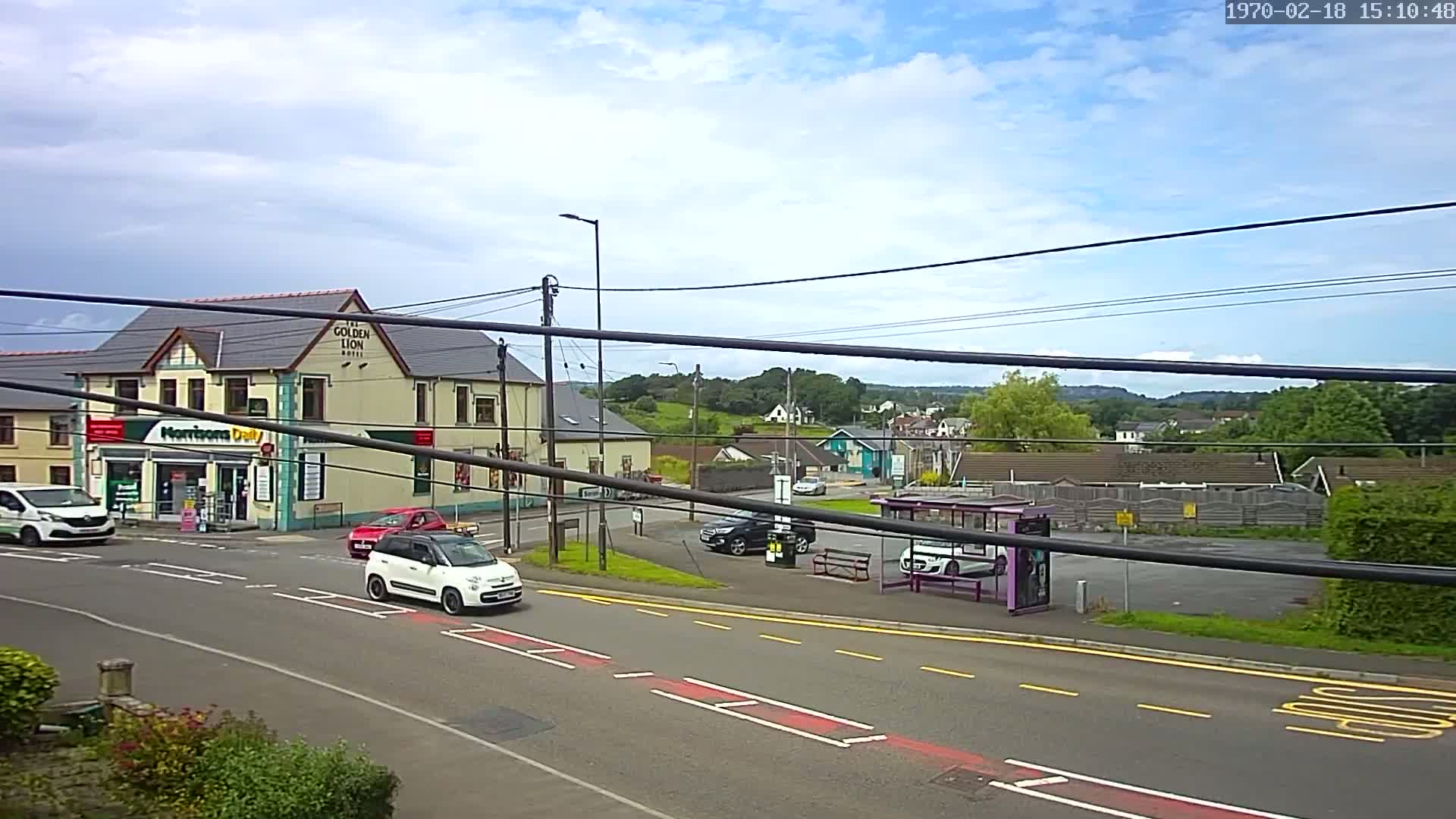 A partly cloudy day reveals a street scene with a few cars, a bus shelter, a small convenience store, and a pub, all set against a backdrop of green hills and houses.