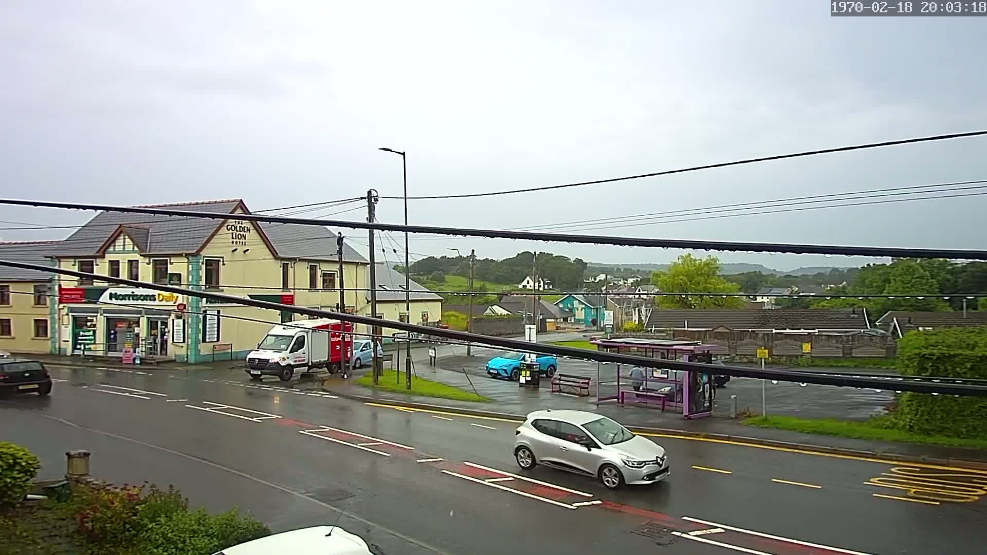 A wet, overcast street scene shows a small town with a pub, a convenience store, several cars, and a bus stop under a rainy sky.