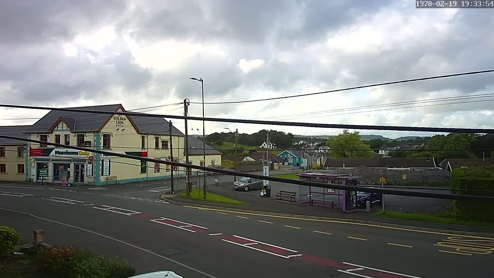 A mostly cloudy day reveals a street scene with a hotel, a small shop, a bus stop, parked cars, and a background of green hills and houses.