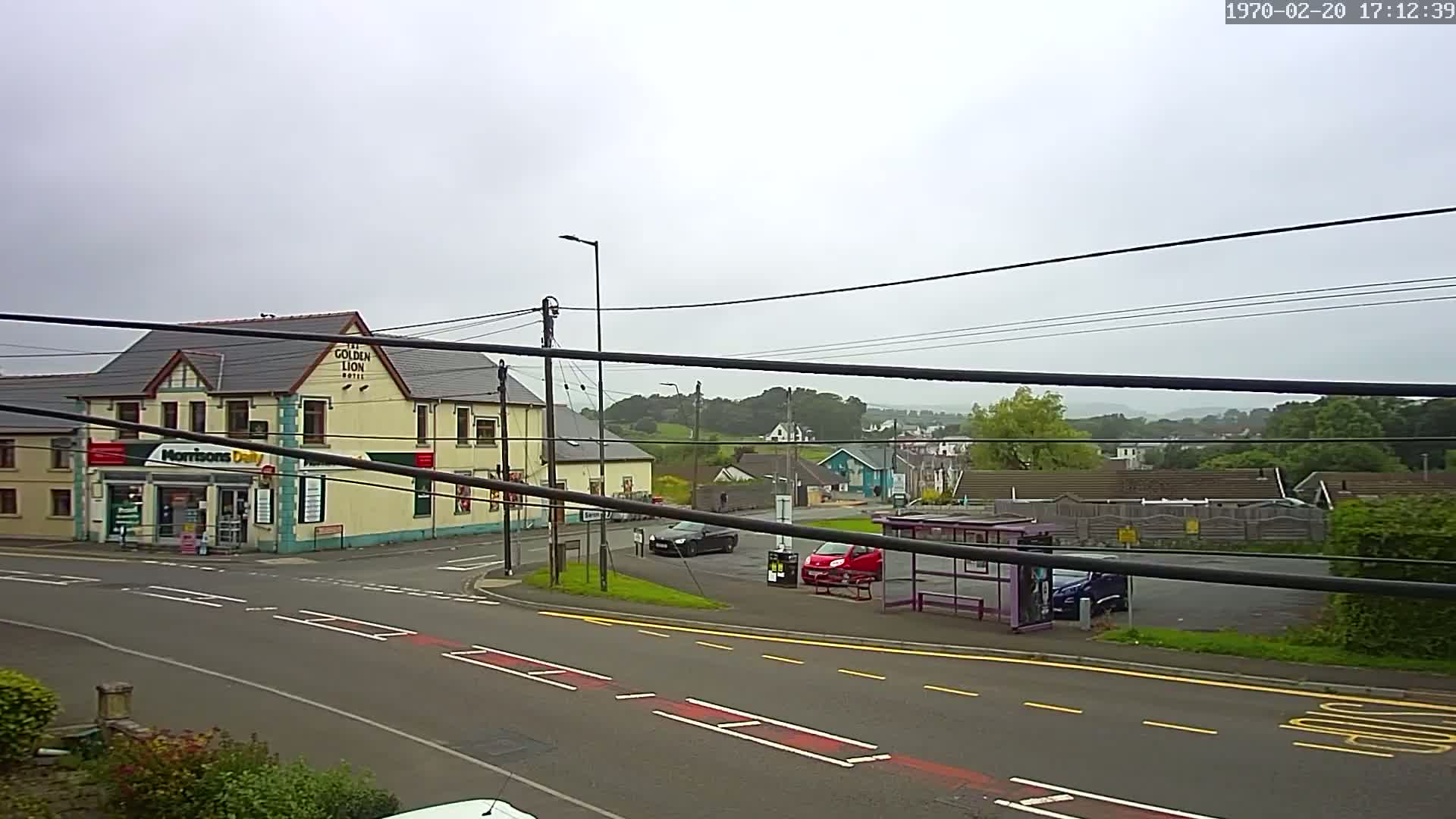 A street scene on an overcast day shows a corner with a hotel, a convenience store, a bus stop, and several cars, all set against a backdrop of rolling green hills.