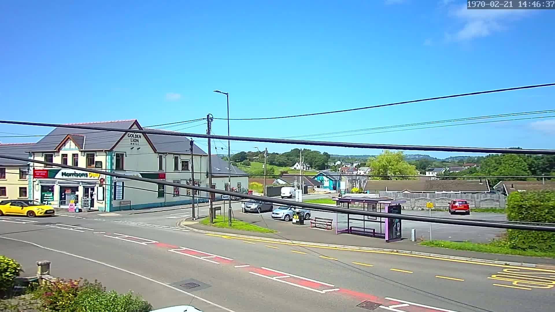 A sunny day reveals a street scene with a two-story building, a bus stop, several cars, and a background of houses and trees.