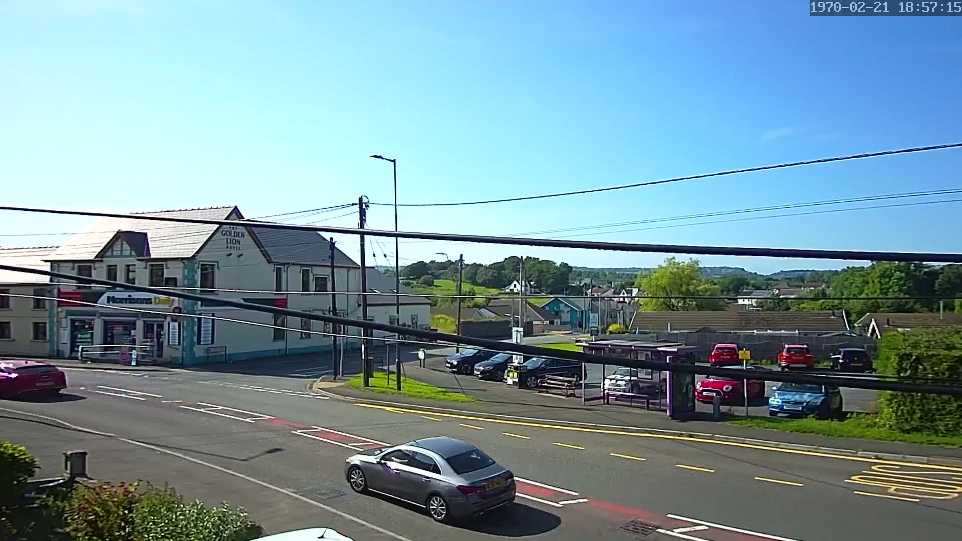 A sunny day reveals a street scene with a building that has a shop on the ground floor and a pub above, several cars parked in a lot, and a bus stop, all situated near a crossroads.