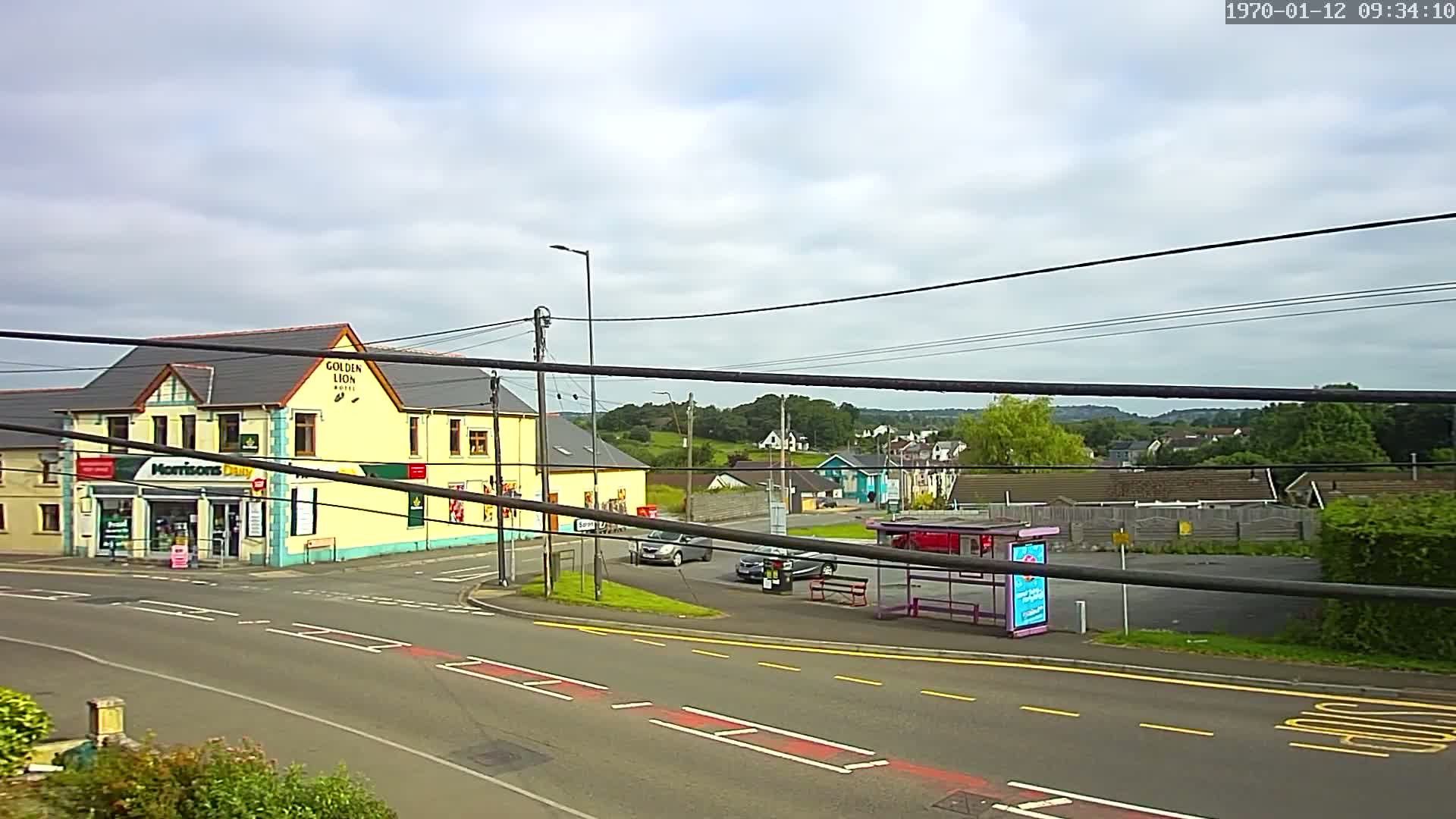 A mostly cloudy day reveals a yellow building with a shop on the ground floor, a bus stop, several cars, and a residential area in the background.