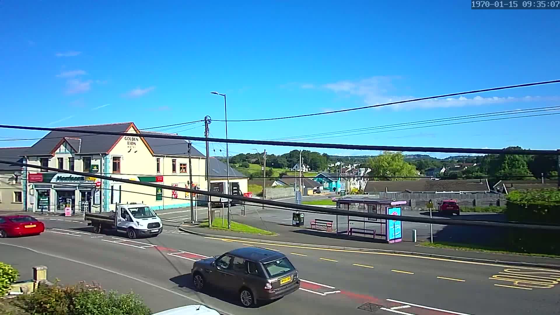 A sunny day reveals a street scene with several cars, a bus shelter, and buildings including a pub and a shop, all set against a backdrop of lush green hills.