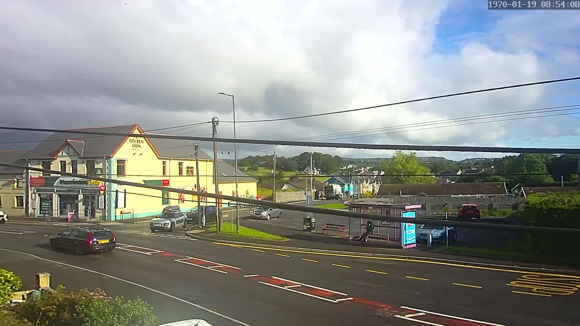 A partly cloudy day reveals a street scene with cars at an intersection near a yellow building, a bus stop, and a background of green hills.