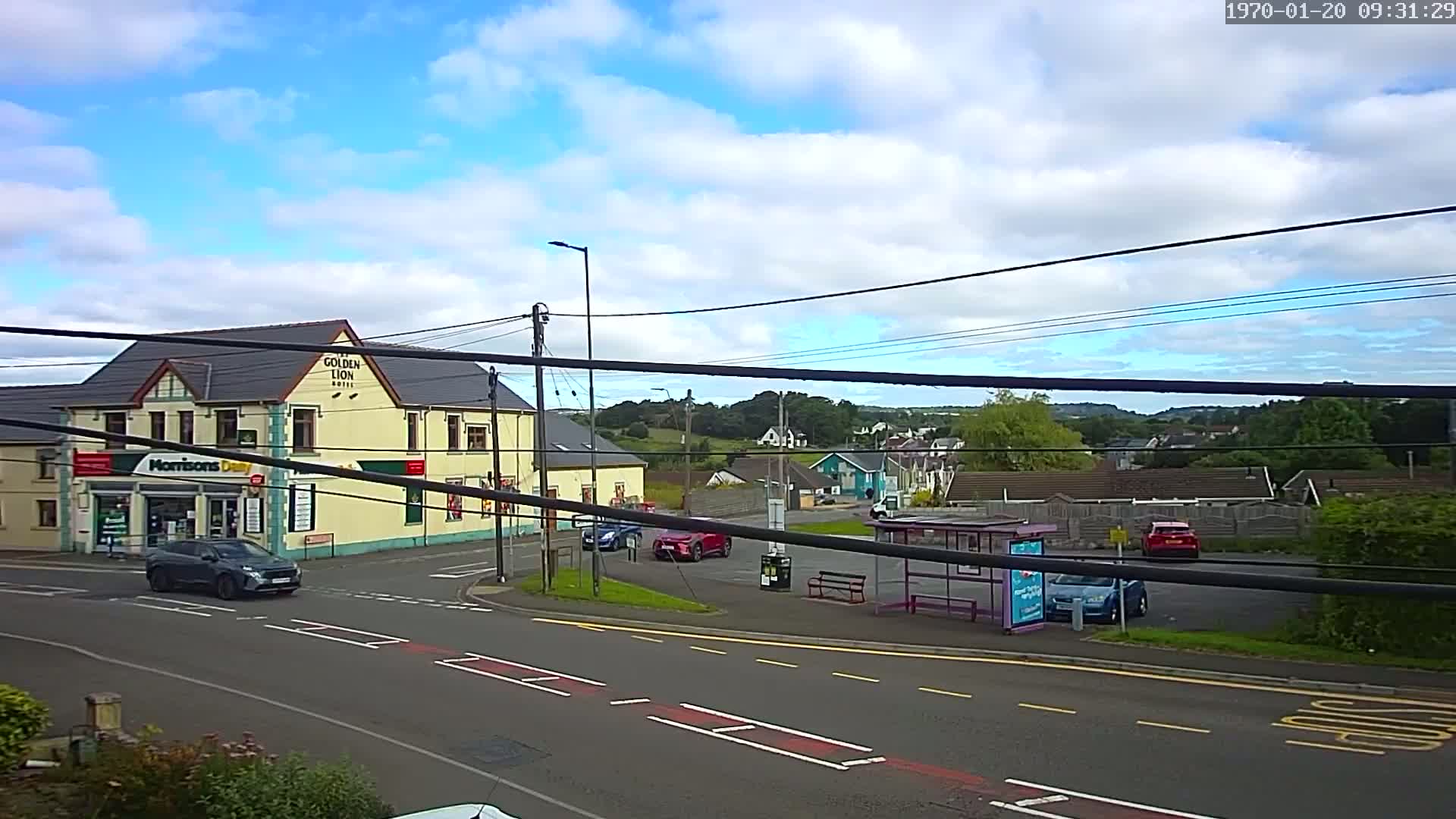 A partly cloudy day reveals a street scene with a hotel, small store, several cars, a bus stop, and residential buildings in the background.