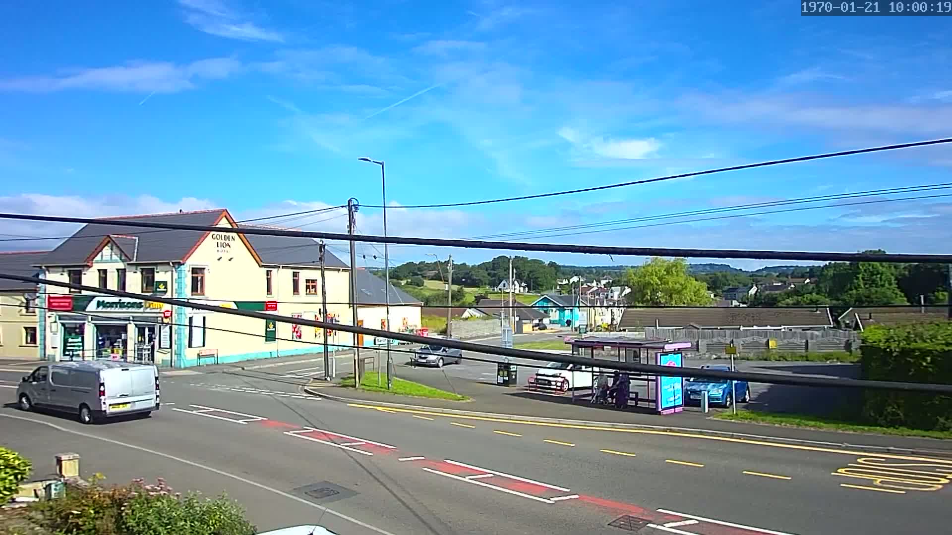 A sunny day reveals a street scene with a pub, a small shop, a bus stop, several cars, and a van, all situated near a tree-lined hill.