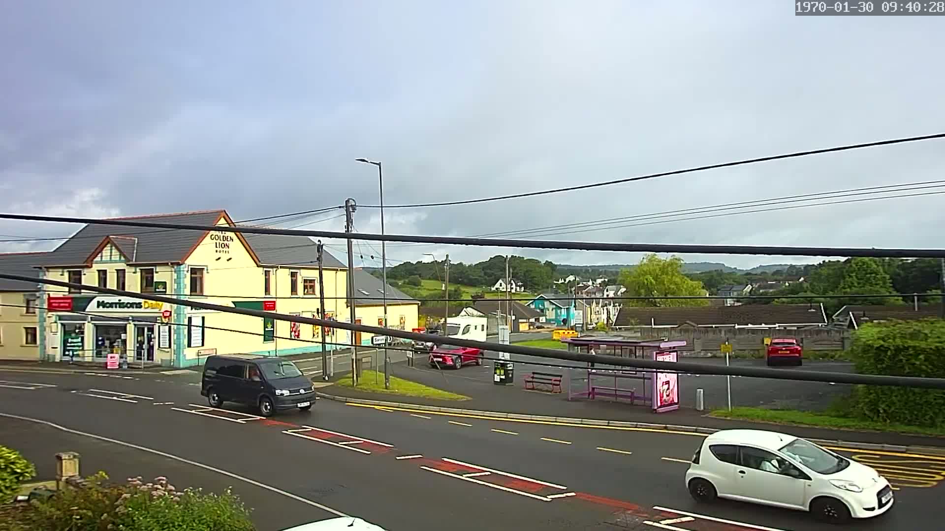 A street scene under an overcast sky shows a yellow building, several vehicles, and a bus stop in a town with green hills in the background.