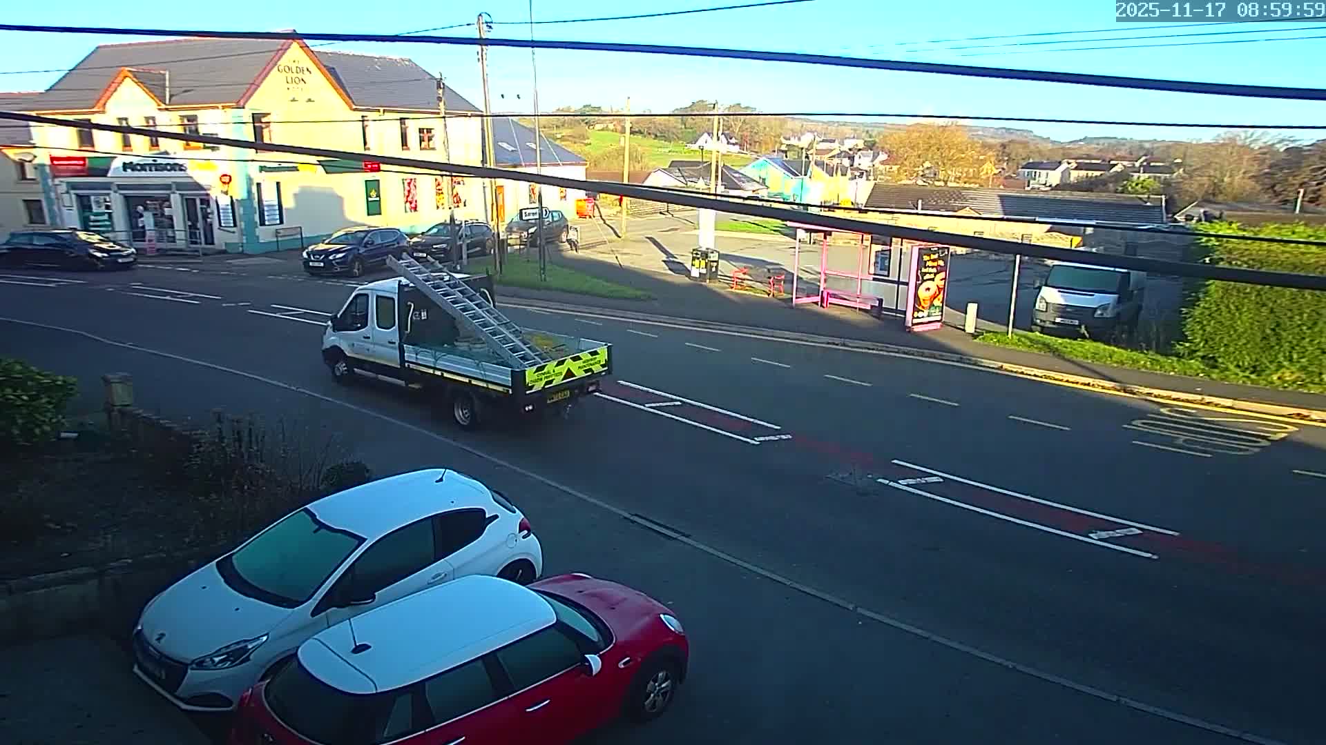 On a clear, cold morning, a white flatbed truck with a ladder drives down a road past a pub, a grocery store, and a pink bus stop, with cars showing frost on their windshields parked in the foreground.