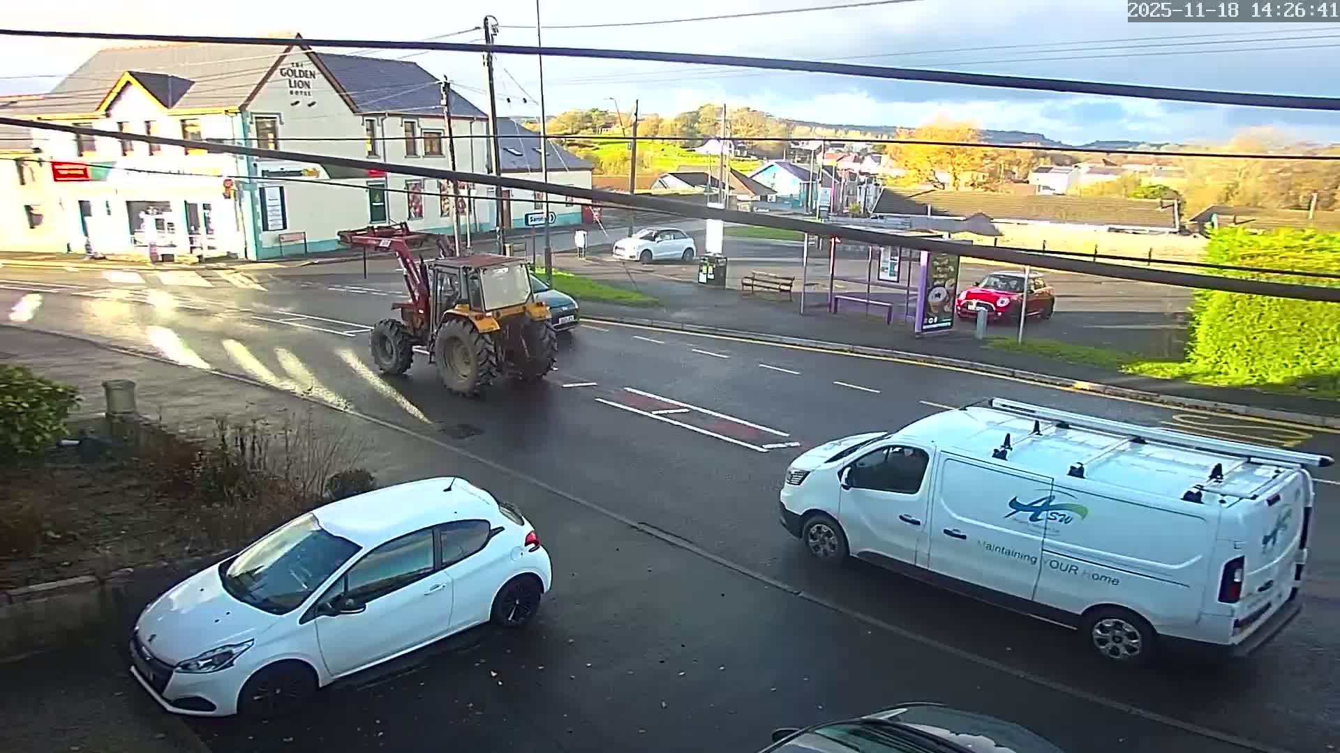 On a clear, cold morning, a white flatbed truck with a ladder drives down a road past a pub, a grocery store, and a pink bus stop, with cars showing frost on their windshields parked in the foreground.
