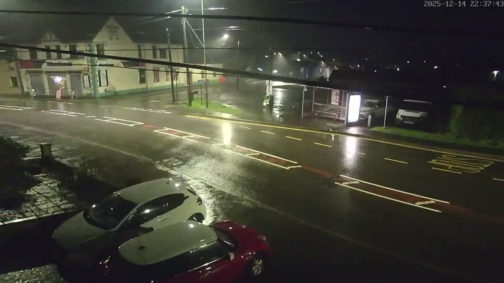 On a clear, cold morning, a white flatbed truck with a ladder drives down a road past a pub, a grocery store, and a pink bus stop, with cars showing frost on their windshields parked in the foreground.
