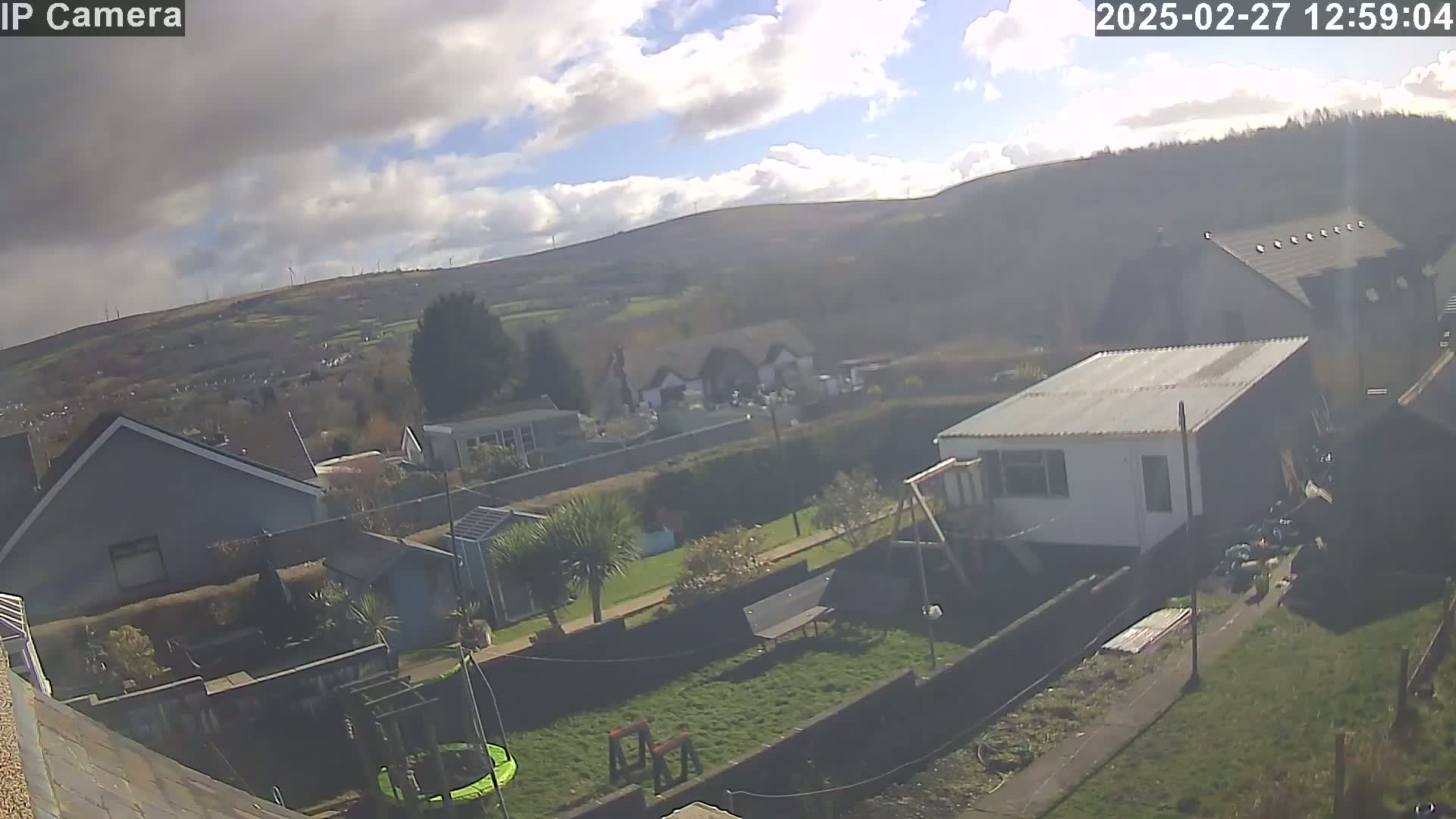 A partly sunny day shows a residential backyard with a trampoline and garden, overlooking a valley with houses and distant hills under a partly cloudy sky.