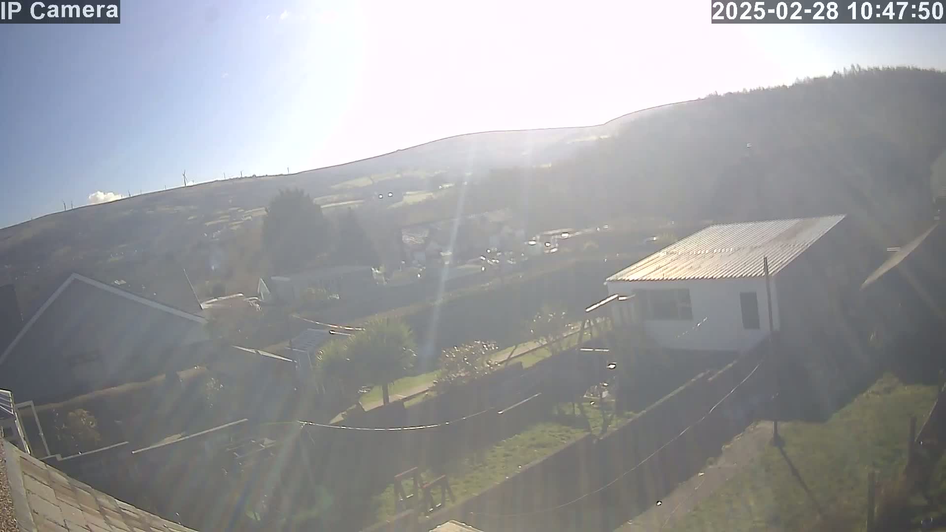 A sunny, hazy view from an elevated position shows a residential area nestled in a valley, with distant hills and wind turbines visible in the background.