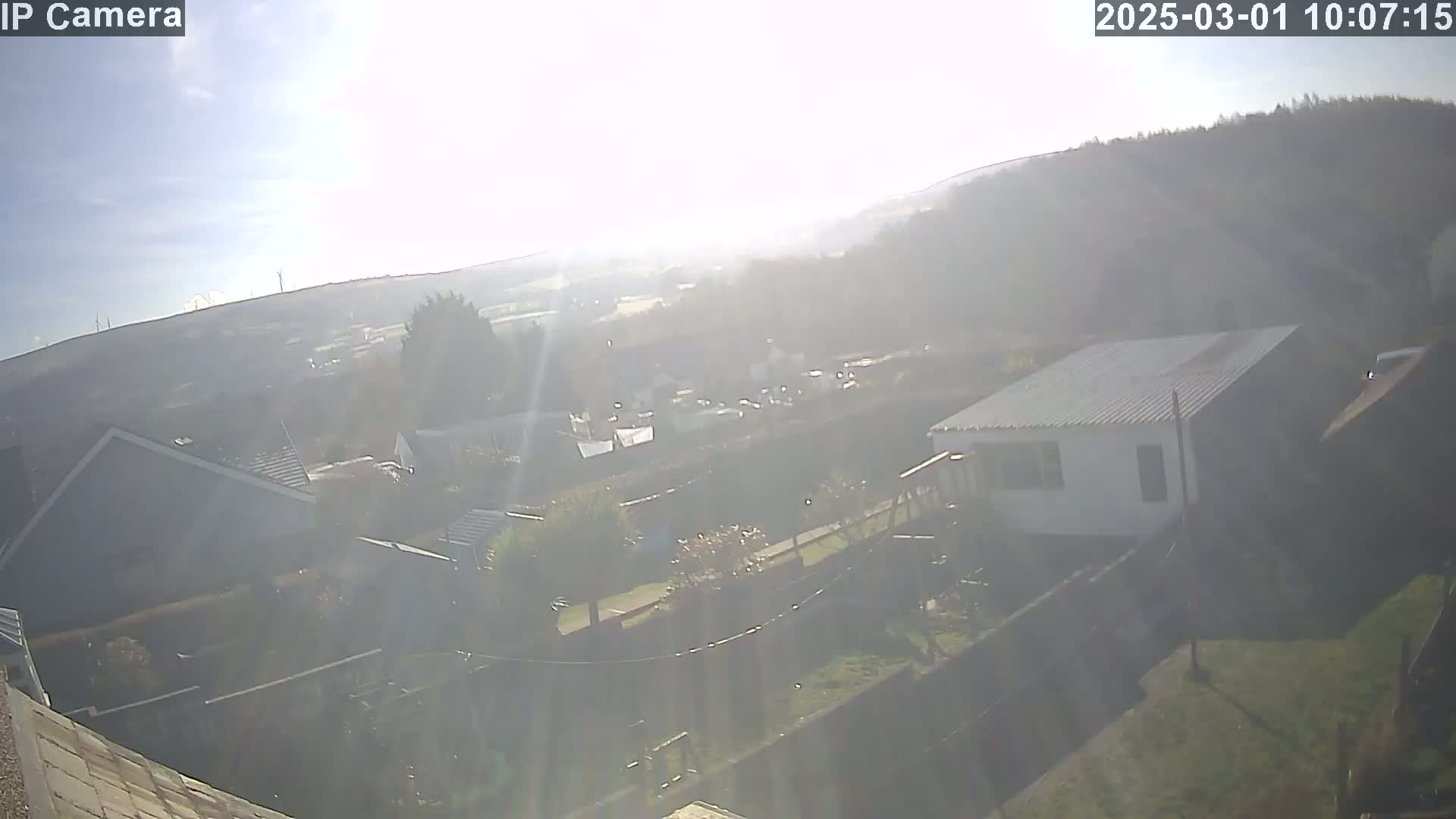 A hazy, sunny day reveals a residential area nestled below a tree-covered hill, with a view extending to distant wind turbines.