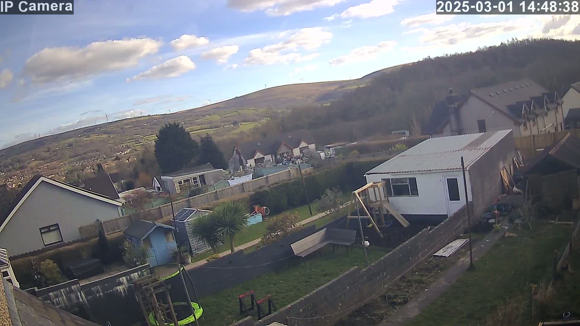 A partly sunny day overlooking a residential area with houses, gardens, and a hilly landscape in the background featuring wind turbines.
