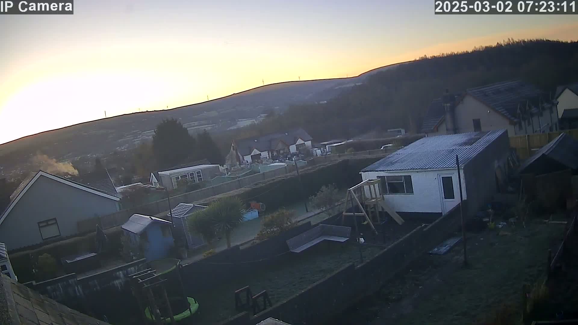 A backyard scene at sunrise shows several houses and a small white shed, with hills visible in the background under a clear, pale sky.