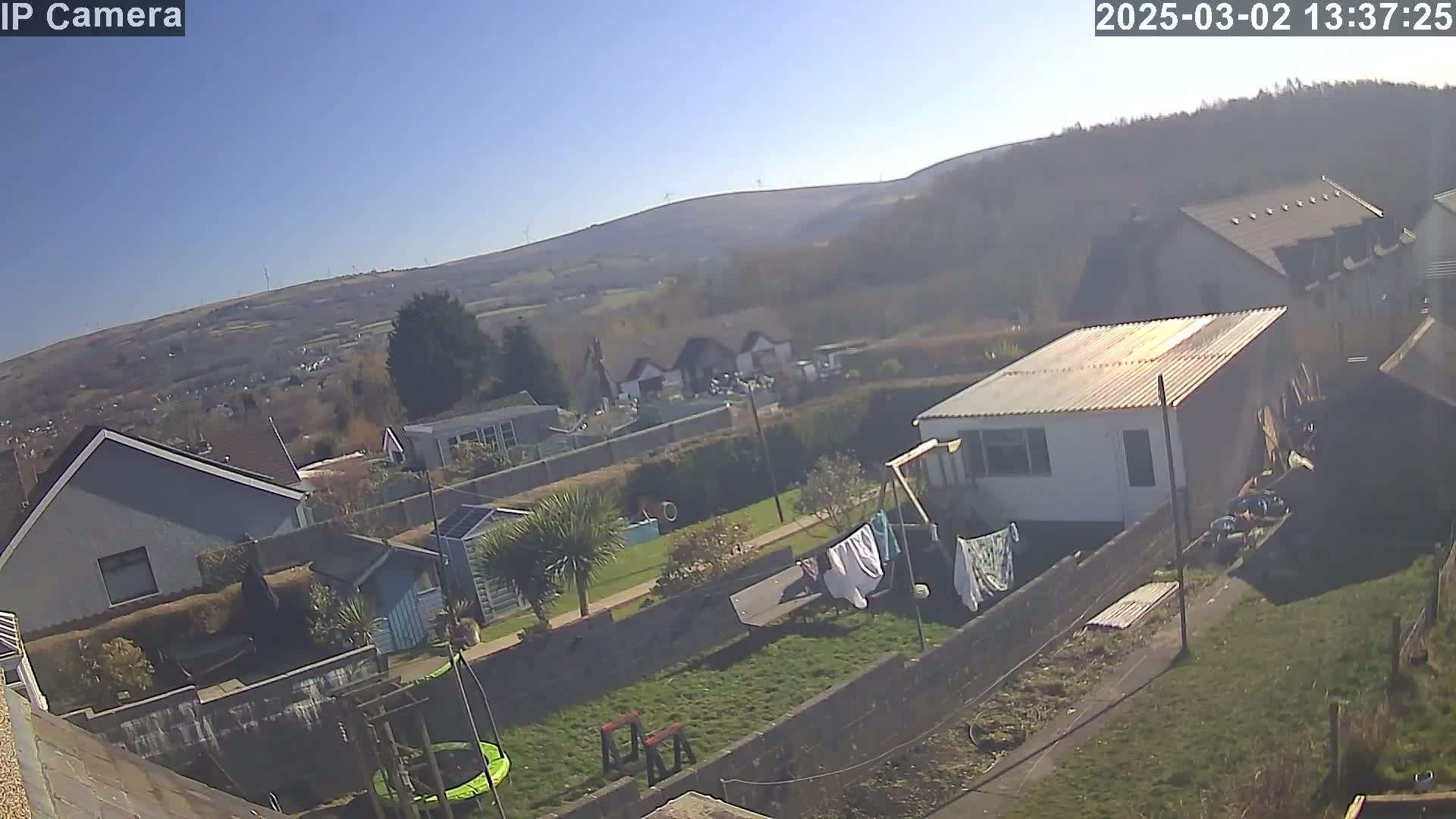A sunny day reveals a backyard scene with houses, a trampoline, and laundry drying in the wind, overlooking a hilly landscape with wind turbines in the distance.
