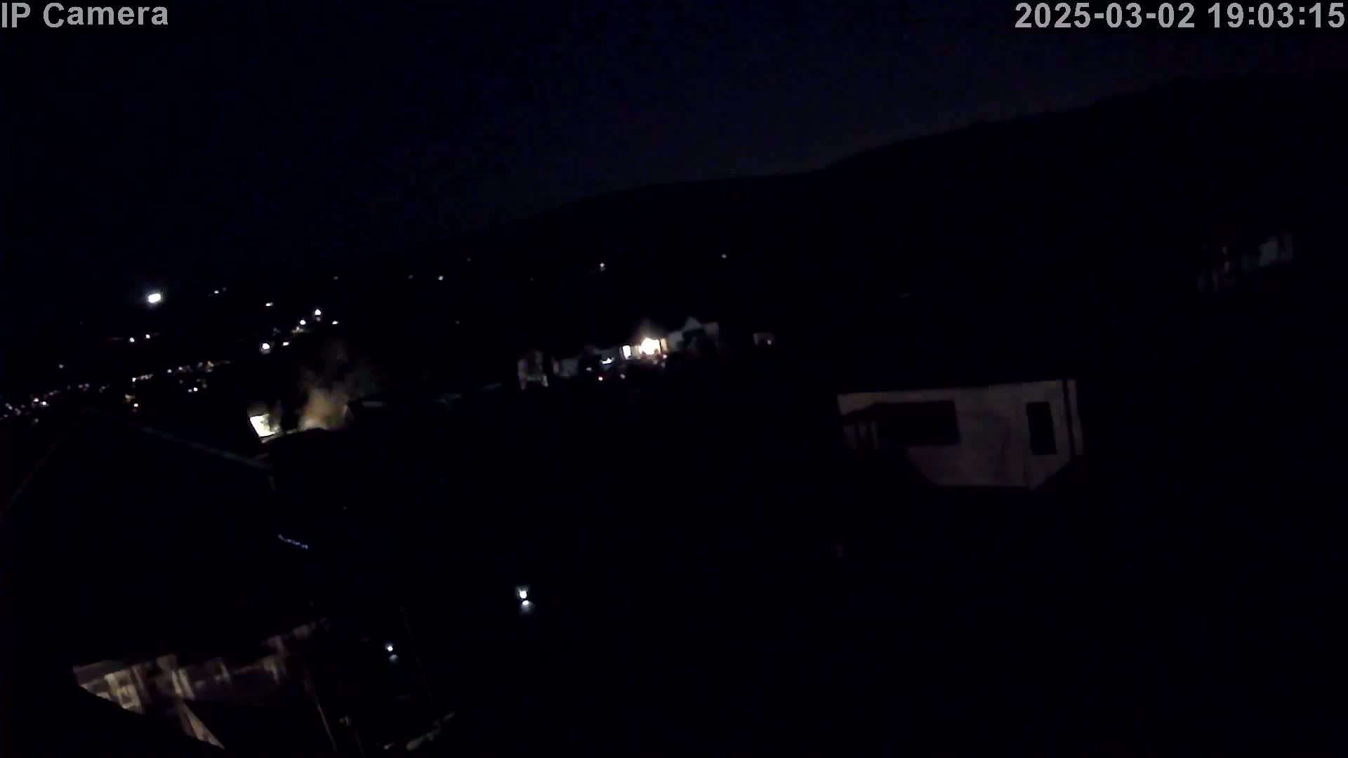 A nighttime, high-angle view shows a dark residential area with sparsely lit houses and a distant hillside under a clear, dark sky.