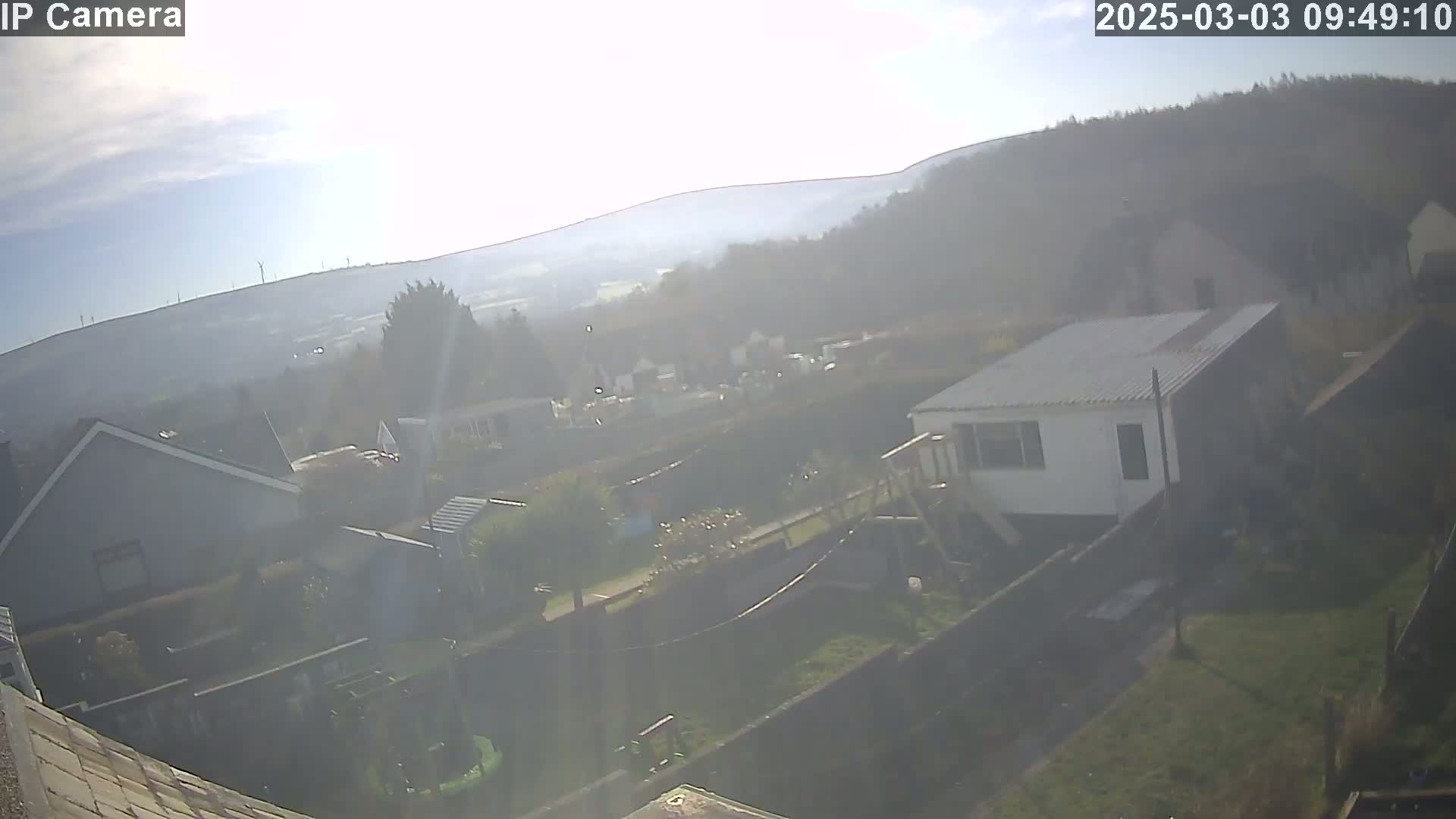 A hazy, sunny day overlooks a residential area with houses, yards, and a distant hillside featuring wind turbines.