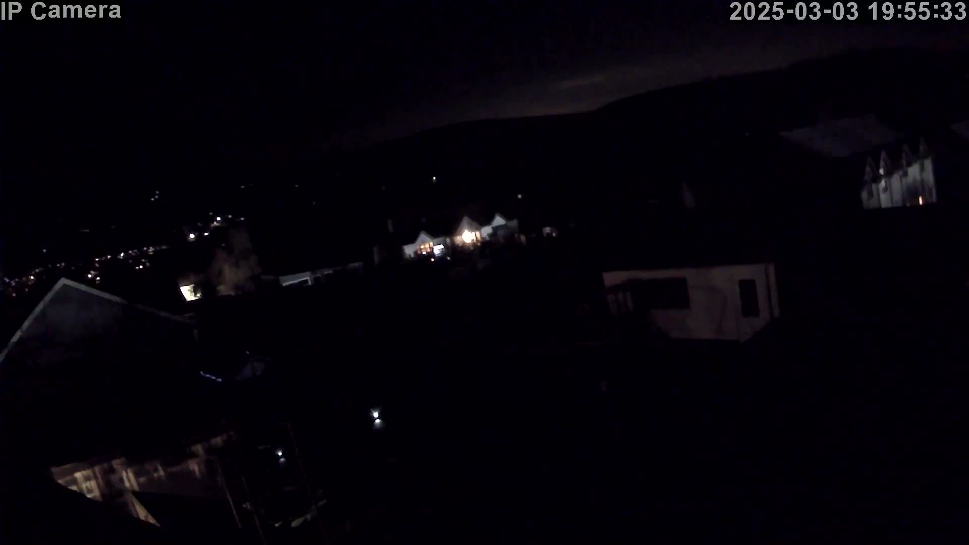 A nighttime, high-angle view shows dimly lit rooftops and houses, with a few interior lights visible, under a dark sky.