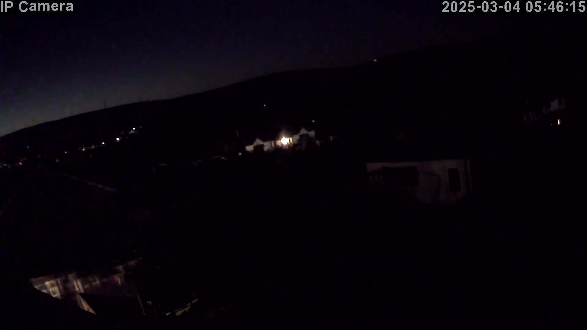 A nighttime, high-angle, long shot shows a dark, residential area with a few dimly lit houses under a clear, dark sky.