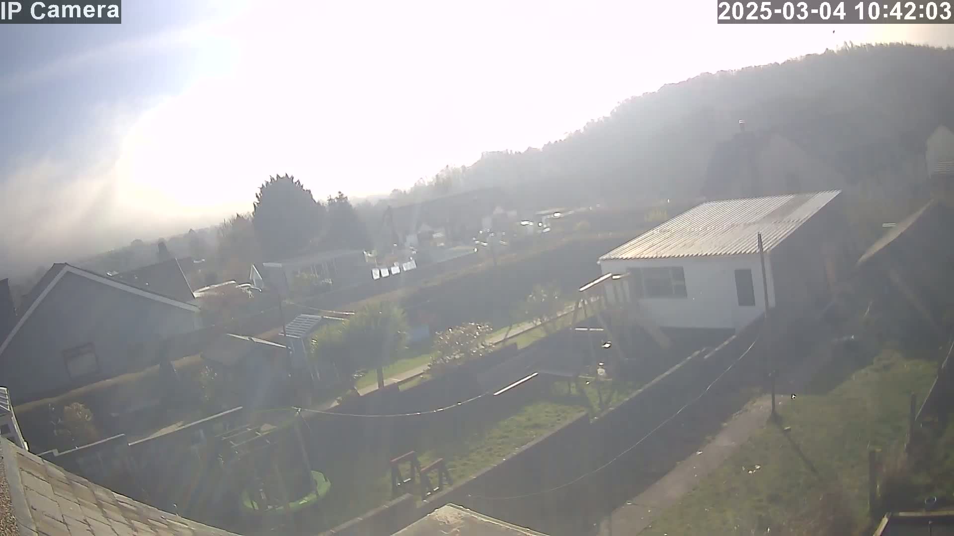 A hazy, sunny day overlooks a suburban backyard with houses and a small outbuilding, including a playset and a partially visible trampoline.