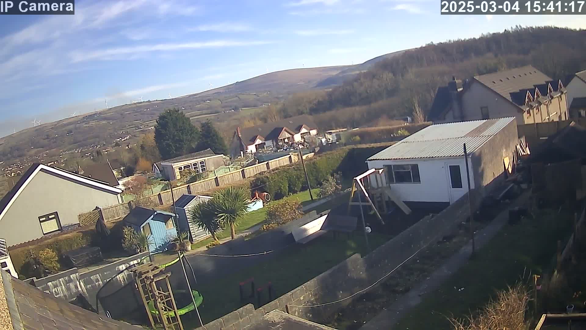 A high-angle, sunny view of a residential backyard with several sheds, a trampoline, and a playset, overlooking a valley with wind turbines in the distance.